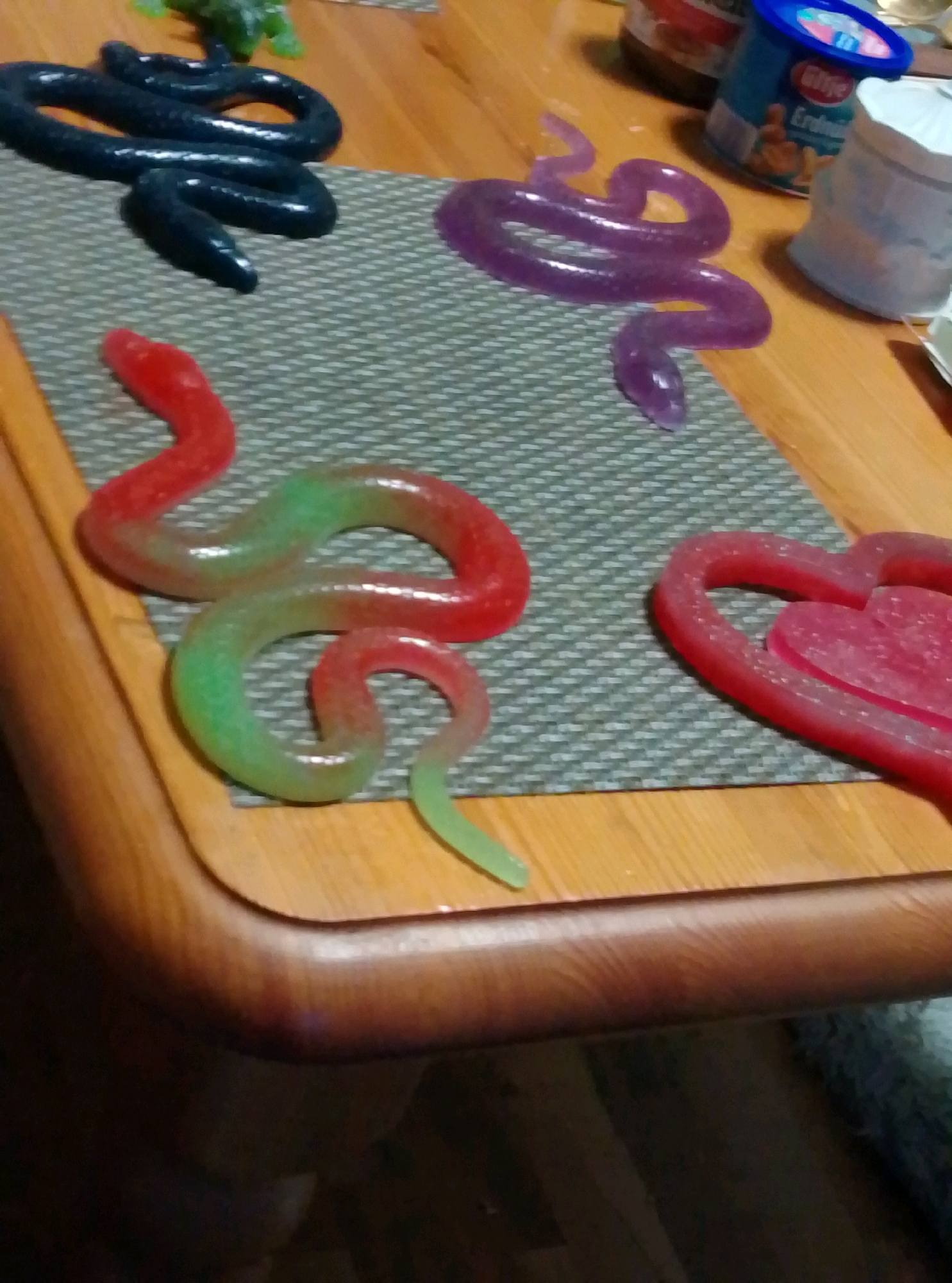 The image displays a collection of gummy candies shaped like snakes and a heart, arranged on a textured placemat on a wooden table. A dark blue gummy snake is coiled in the upper left quadrant. To its right, a translucent purple gummy snake is also coiled. In the lower left quadrant, a red and green gradient gummy snake is partially visible. To the right of this snake, a red heart-shaped gummy candy is present. In the background, on the wooden table, are two containers, one appearing to be a jar and the other a blue plastic tub with a label. The overall lighting suggests an indoor setting. The image does not contain any information related to the city of Wedel, Germany.