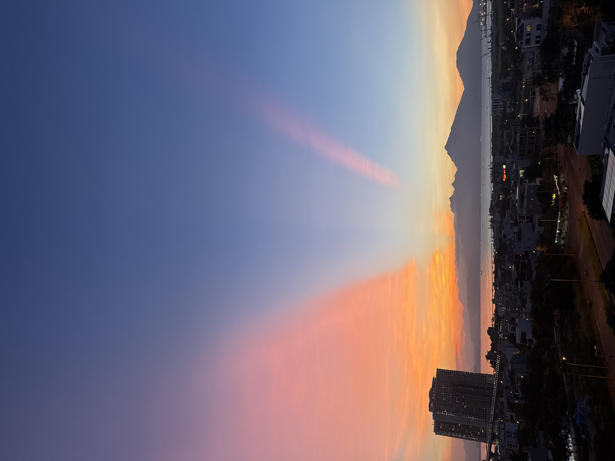 The image captures a sunset over Đà Nẵng, Vietnam. The sky transitions from a deep blue to orange and pink hues near the horizon. Streaks of pink clouds are visible in the upper sky. In the distance, a mountain range divides the sky from the city. Buildings and streetlights illuminate the urban landscape, with a tall building prominently featured in the foreground. The body of water and the port area are faintly visible in the middle ground. A road with a few streetlights is visible in the right foreground.