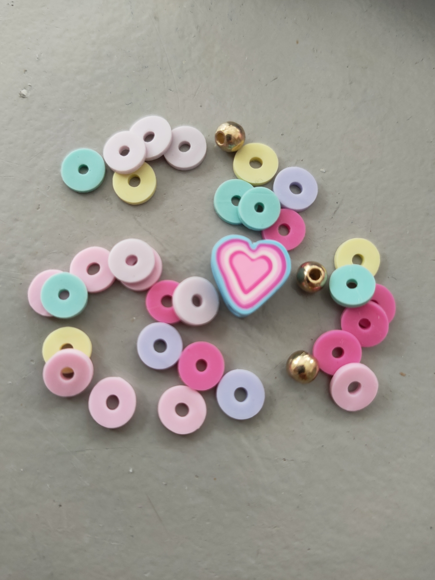 The image displays a collection of colorful, flat, round beads and three gold beads scattered on a gray, textured surface. A single heart-shaped bead, composed of concentric rings of blue, white, and pink, is centrally located among the other beads. The lighting suggests an indoor setting, possibly a table or floor, with the light source appearing to be overhead. There is no indication of a specific location within the city of Perpignan, France. The scene is static and appears to be a close-up shot of craft supplies.