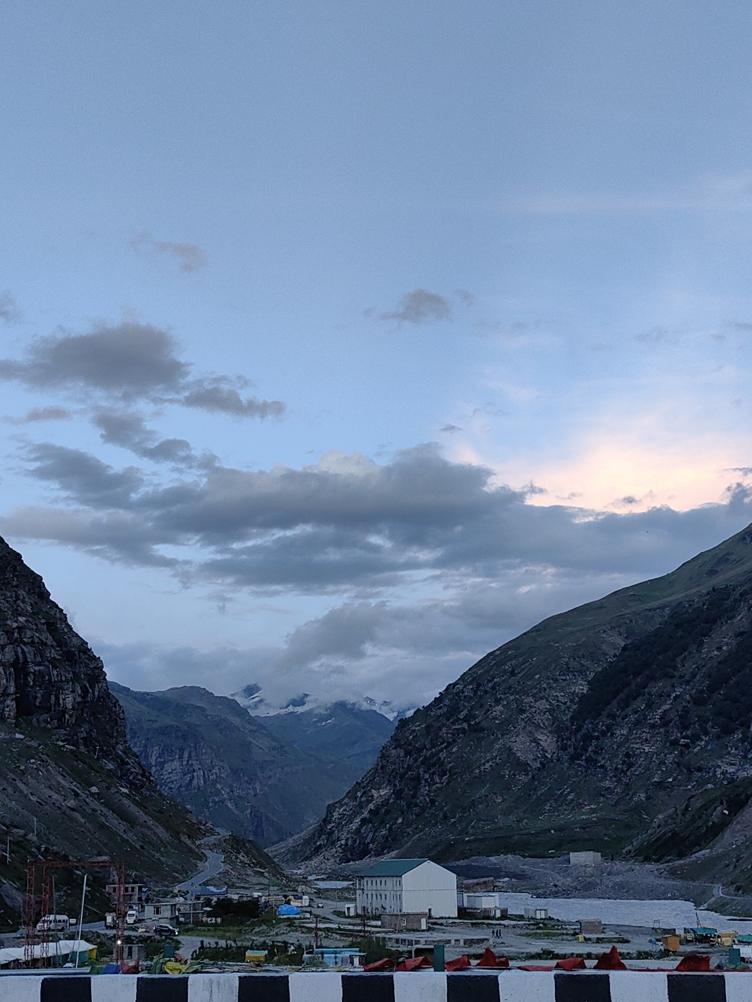 An exterior, vertical, full shot captures a valley landscape at dusk. The sky is a pale blue with scattered clouds, some of which are tinged with pink from the setting sun. On either side of the valley, steep, rocky mountains rise up, covered in patches of green vegetation. In the center of the valley, a road winds through a small settlement. Buildings, some appearing to be under construction, are visible, along with several vehicles. The overall scene conveys a sense of remoteness and natural beauty.