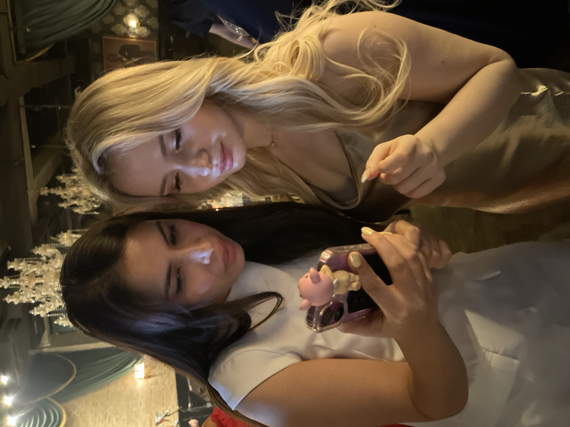 The media file shows two young women, one with long blonde hair and the other with long dark hair, in a dimly lit indoor setting. The blonde-haired woman is wearing a shiny, champagne-colored dress and is leaning forward, looking down towards the other woman. The dark-haired woman is wearing a white top and is holding a smartphone with a pink case adorned with a small pig figurine. She is looking at the phone. The background appears to be a restaurant or bar with decorative elements like curtains and chandeliers, suggesting an evening event. The lighting is warm and atmospheric, indicative of nighttime. The women's expressions suggest a casual, friendly interaction. There is no visible text in the image. The context of South Jakarta, Indonesia, implies an urban environment.