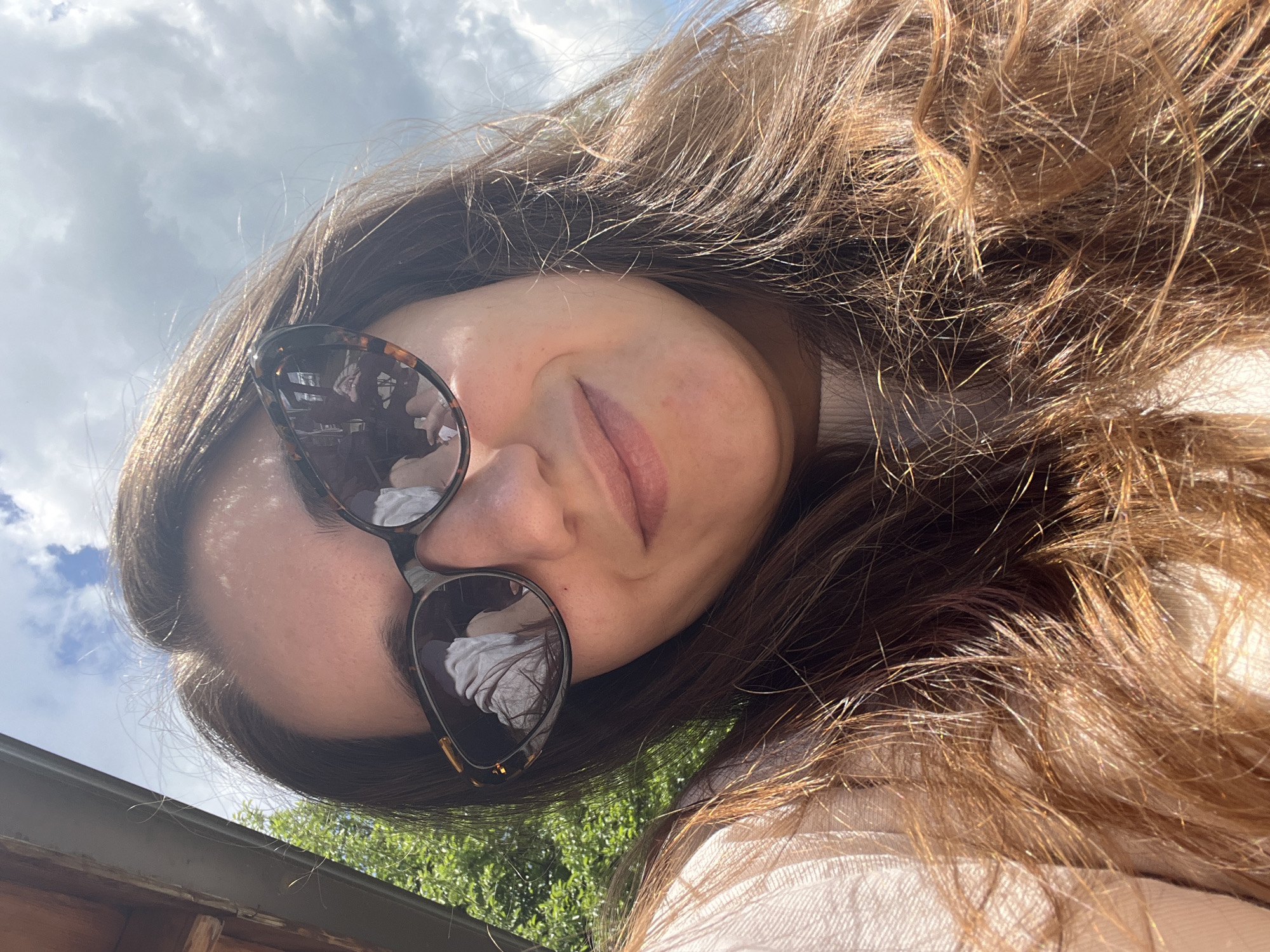 A selfie image shows a woman with brown hair and sunglasses. The reflection in the sunglasses shows an interior space with white fabric. The background consists of a partly cloudy sky and green foliage. A wooden structure is also visible in the lower part of the image. The picture likely taken outdoors, location in the city of Gorno Kamartsi, Bulgaria cannot be confirmed from the image.