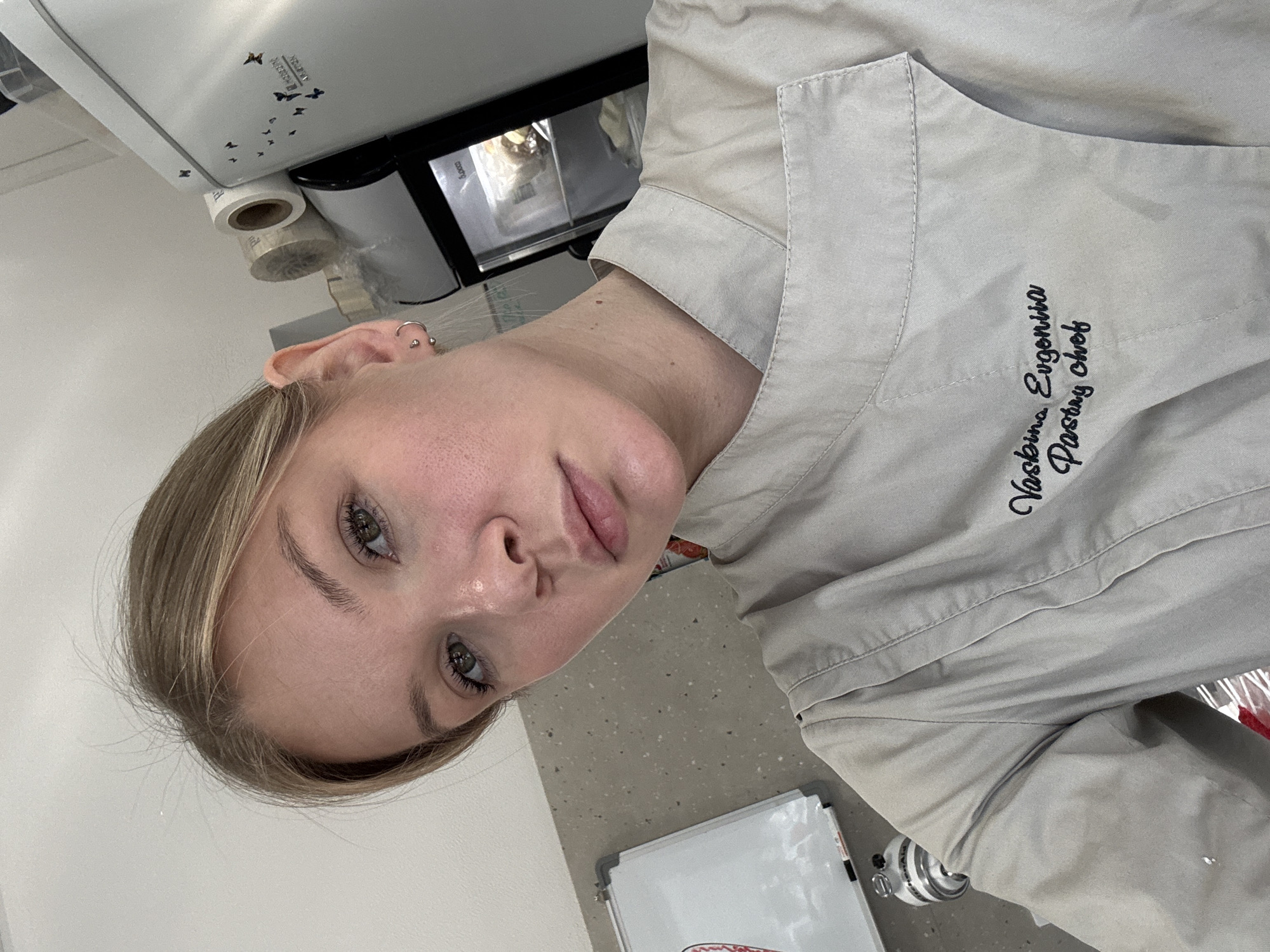 A medium, angled, indoor shot shows a woman with blond hair and green eyes wearing a chef's coat with the name "Vaskina Evgeniia Pastry Chef" embroidered on it. The woman is facing the camera with a slight tilt of her head to the right. In the background, there is a white wall, a machine with a roll of paper on it, and a small refrigerator. A whiteboard is visible in the lower right corner of the frame. It is likely that this photograph was taken in Perm, Russia.