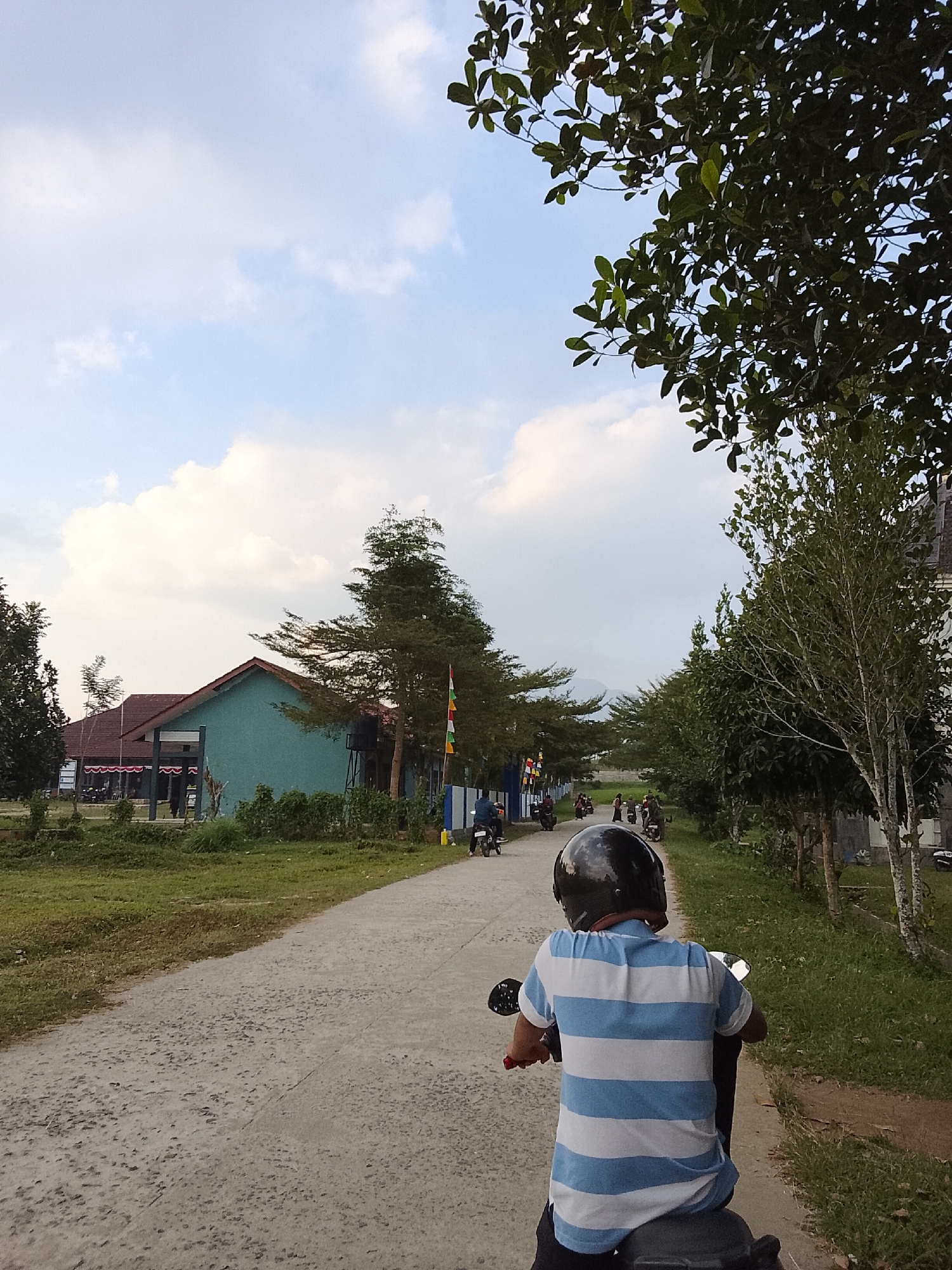 The image depicts an outdoor scene on a paved road, likely in Padang Dalom, Indonesia, under a cloudy sky with some blue patches visible. In the foreground, a person wearing a black helmet and a blue and white striped t-shirt is riding a motorcycle. The road stretches into the distance, lined with trees and some buildings. On the left side of the road, a teal-colored building with a brown roof is visible, possibly a school or community center, with flags and people near it. Several other motorcycles are seen on the road further ahead, indicating ongoing activity. The environment appears to be a semi-rural or suburban area, with vegetation and a generally calm atmosphere.