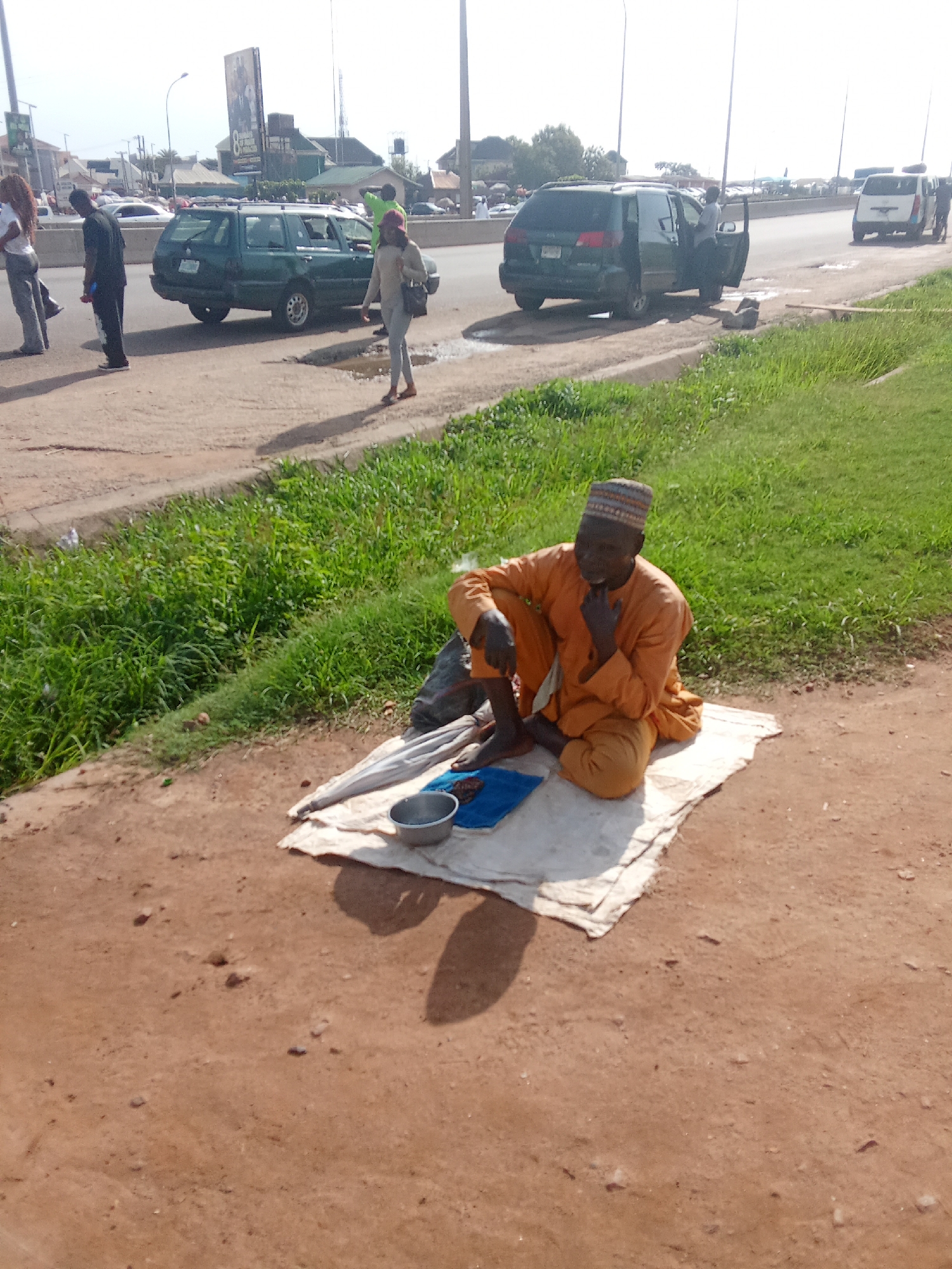 The image shows a man sitting on a white cloth on the ground, in front of a grassy embankment. He is wearing an orange outfit and a patterned cap. Next to him are a silver bowl and a folded umbrella. In the background, a busy road with cars and other people is visible. The scene appears to be in an urban environment, possibly along the side of a highway or main road. The lighting suggests it is daytime, with a bright sky.