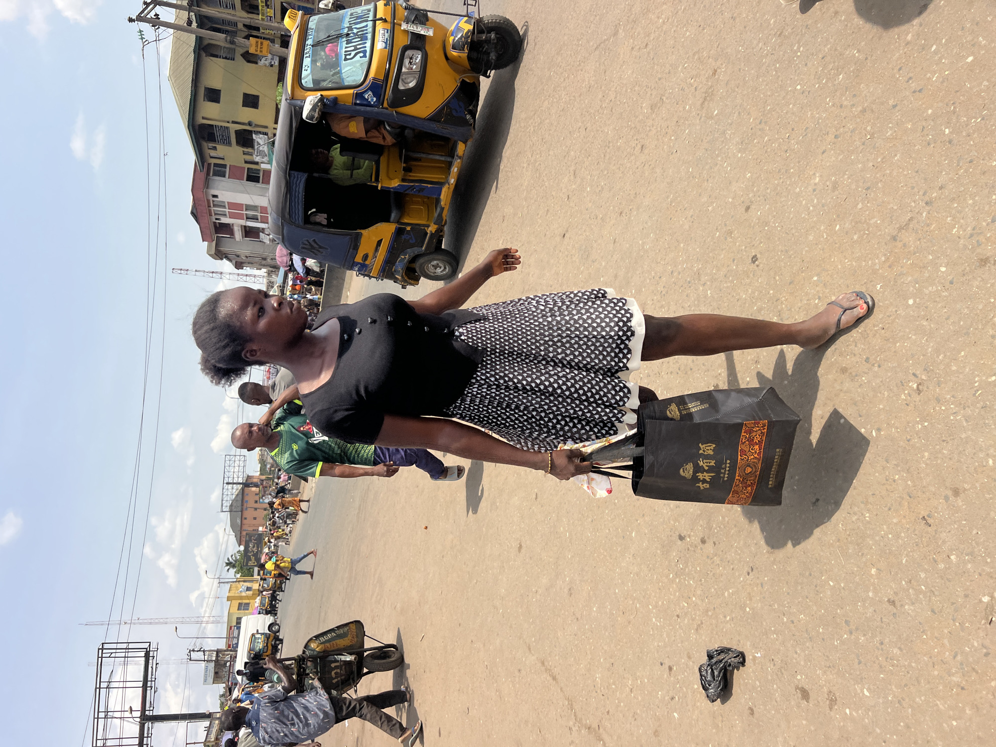 A street scene in Onitsha, Nigeria. A young woman wearing a black top and a white and black patterned skirt walks across the street, carrying a black shopping bag with Chinese characters. She is wearing flip-flops. To her left, a yellow auto-rickshaw is parked. Other people are walking in the background. A man wearing a green shirt is walking past her. A person is pushing a cart with items on the left side of the image. The street is paved and dusty. Buildings and power lines are visible in the background. The sky is partly cloudy.