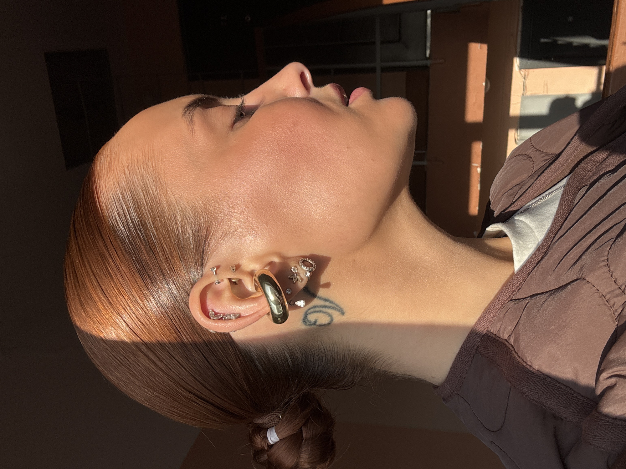 A young woman with light skin and auburn hair is shown in a close-up, angled view of her face and upper body. Her hair is styled in a slicked-back fashion and gathered in a braid at the nape of her neck. She is wearing multiple earrings in her left ear, including a large gold hoop and several smaller studs and rings. A small tattoo is visible on her neck just below her ear. She is wearing a brown quilted jacket over a white top. The lighting is strong and directional, creating sharp contrasts of light and shadow on her face, hair, and clothing. The background is blurred and out of focus, but suggests an interior setting with a window or doorway visible. There is no information to indicate that the photo was taken in the city of Chaikovsky, Russia.
