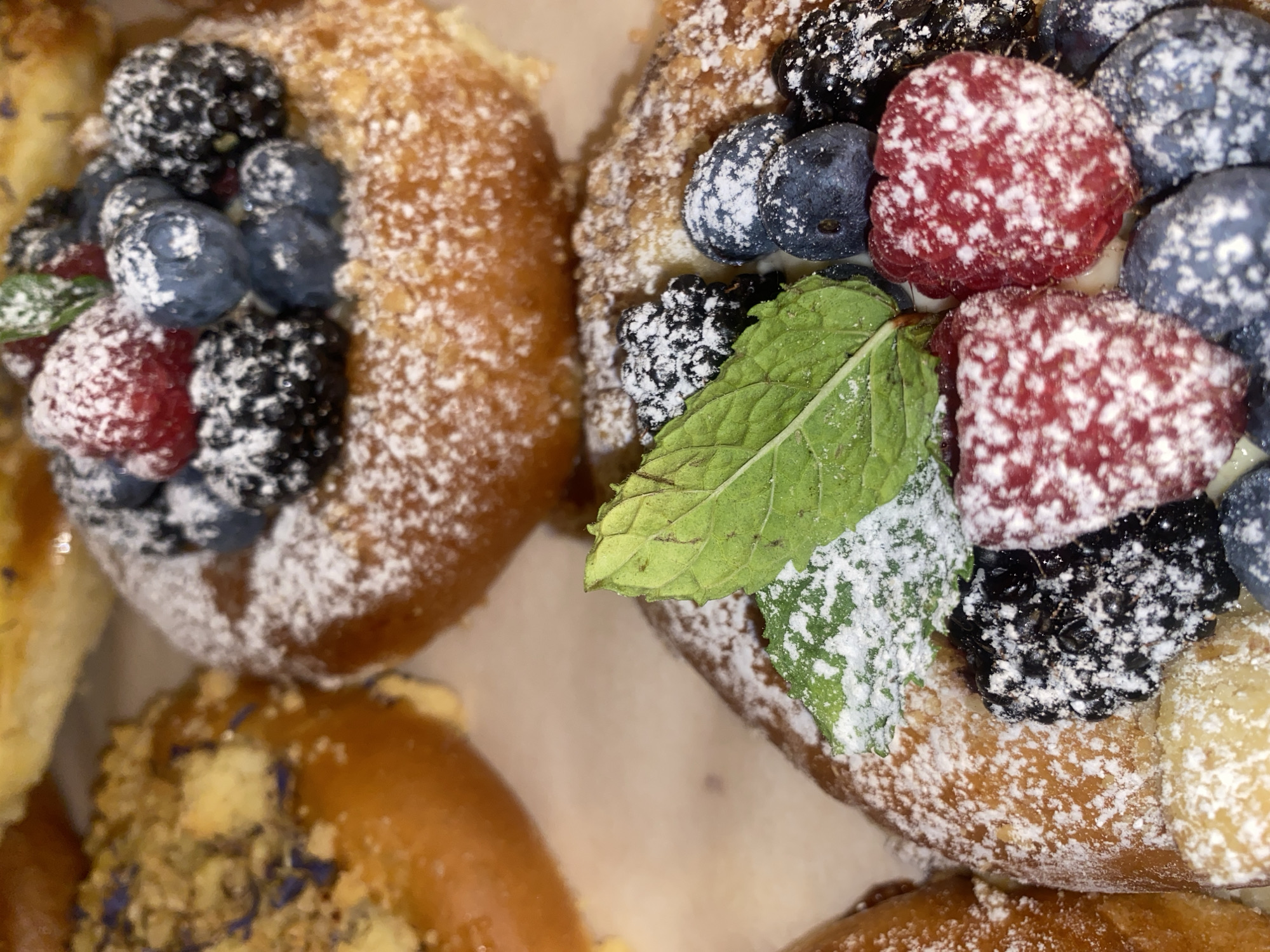 The image is a close-up, overhead shot of several pastries, likely served at a bakery or cafe. The pastries are golden brown and topped with fresh berries such as raspberries, blueberries, and blackberries. They are dusted with powdered sugar and some have a sprig of mint. The background is a light-colored surface, possibly a table or serving platter. There are no people or indications of location within the city of Warsaw, Poland.