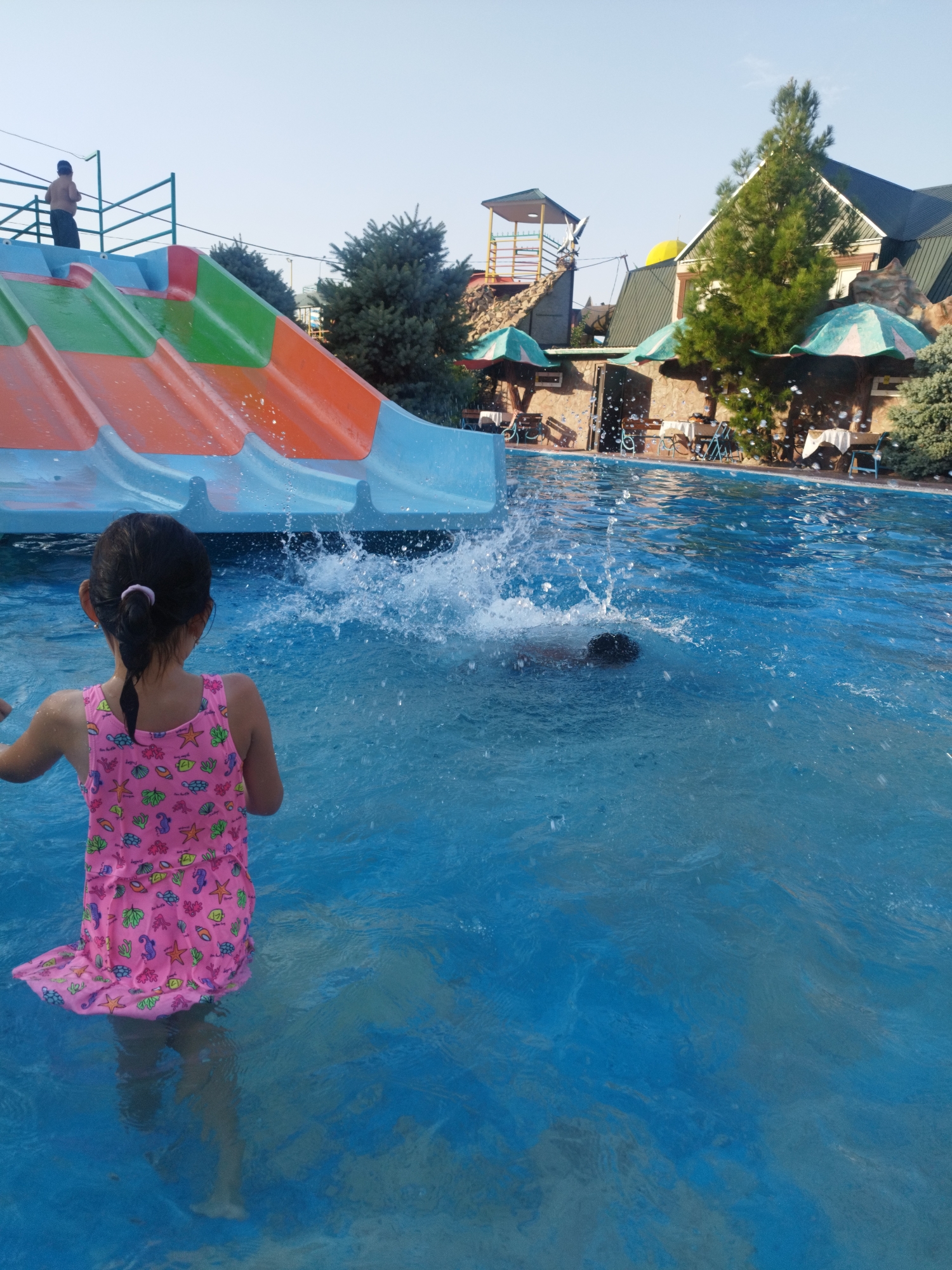 At a water park in Shymkent, Kazakhstan, a young girl in a pink swimsuit with a shell pattern stands in a bright blue swimming pool. She is facing away from the camera, looking towards a water slide. To her right, another person is submerged in the water, with only their head visible above the surface, creating a splash. The water slide, predominantly orange and green with blue accents, arcs into the pool. In the background, a two-story building with a dark green roof and awnings provides shade over outdoor seating areas with tables and chairs. Evergreen trees and shrubs are scattered throughout the scene, and the sky is clear and bright.