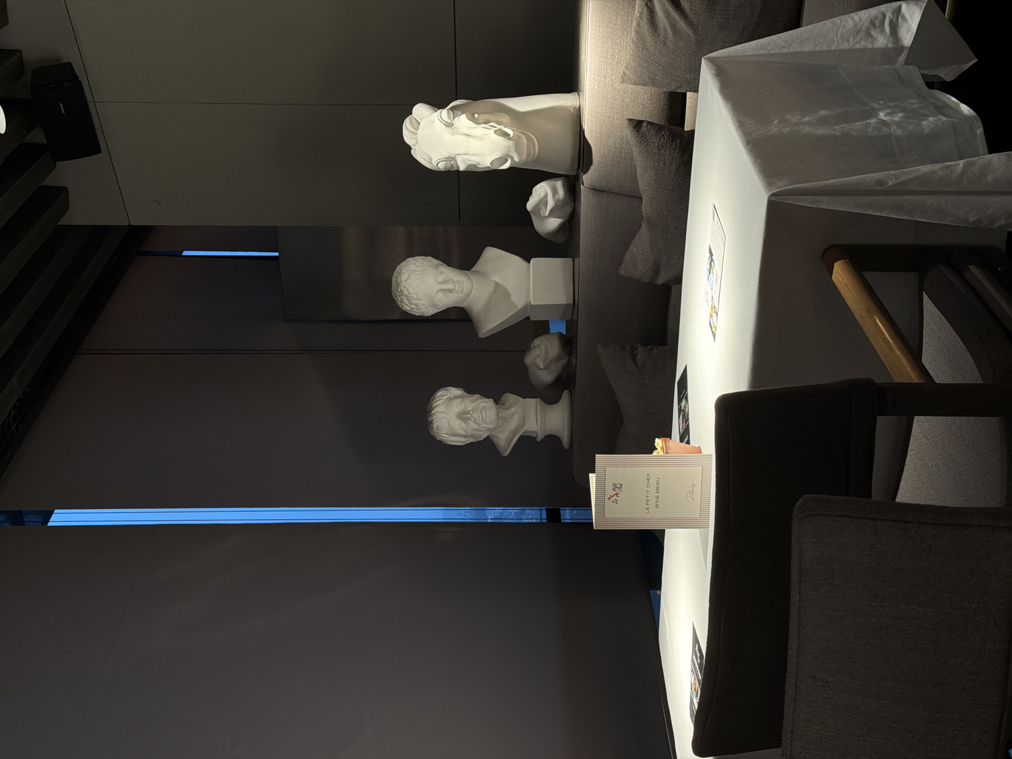The image displays a dimly lit, sophisticated indoor setting, presented sideways with the bottom of the room towards the right. It appears to be a modern restaurant or lounge, characterized by a blend of dark tones, classical artistic elements, and contemporary lighting.
On the left side of the image, which represents a dark wall or display area, several white classical sculptures are arranged. These include a prominent bust of a man, a horse's head, and smaller, rounded forms, all facing towards the top of the image. A striking, narrow blue LED light strip runs horizontally across this dark surface, adding a vibrant modern touch. In the upper left corner, a dark, angular structure, possibly part of a staircase or tiered seating, is partially visible.
On the right side, a dining table is covered with a crisp white tablecloth, suggesting readiness for guests. Beside the table are dark, upholstered chairs, one of which clearly shows a wooden armrest. Behind the table and chairs, a comfortable-looking grey sofa or banquette with dark cushions provides additional seating.
A menu stands upright on the white tablecloth. Clearly legible text on the menu reads "LE PETIT CHEF" and "WINE MENU". The word "Paris" is visible at the bottom, and a logo at the top appears to be "Hôtel La Cité".
The overall atmosphere is quiet and elegant, with no people visible in the scene. The dim, intentional lighting and the glowing blue strip suggest the photo was taken during the evening or night. While the contextual location is Tbilisi, Georgia, there are no specific visual cues within the image itself to indicate this.