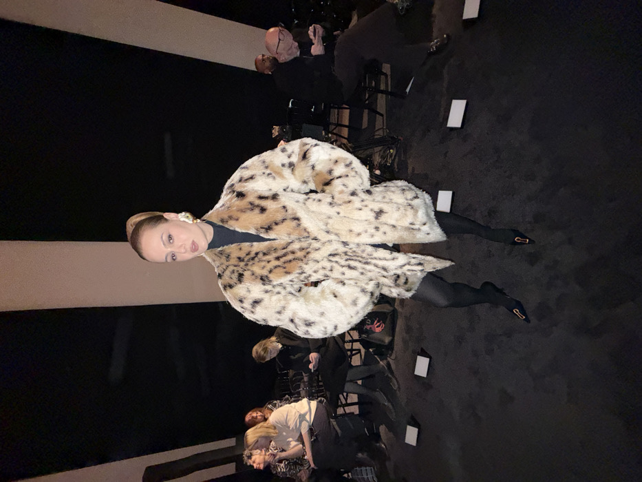 This image captures an indoor scene at a fashion event, likely in Paris, France. A woman stands centrally in the foreground, facing the viewer directly. She wears a voluminous faux fur coat featuring a cream and dark brown leopard print, a black turtleneck, and black opaque tights. Her feet are clad in black pointed-toe heels with gold-toned buckle embellishments. She has light-toned skin, her hair is pulled back in a high ponytail, and she wears large, ornate gold-colored earrings. Her expression is neutral, with slightly parted lips.
Behind the central figure, an audience is seated in rows on dark chairs. On the left side of the frame, multiple individuals are visible, including a woman with light brown hair wearing a light-colored top, and another with reddish-brown hair. A dark bag featuring a red squid-like graphic rests on the floor beside one of these seated individuals. On the right side, a bald man with glasses is visible, engrossed in a mobile phone, with another individual partially obscured behind him. The floor is dark, possibly carpeted, and features several small, white rectangular markers positioned in front of the audience seating. The background consists of large, dark, angled architectural or structural elements with contrasting lighter-colored vertical lines, suggesting the interior of a modern venue or temporary structure. The lighting is focused, illuminating the central subject against a dimmer background.