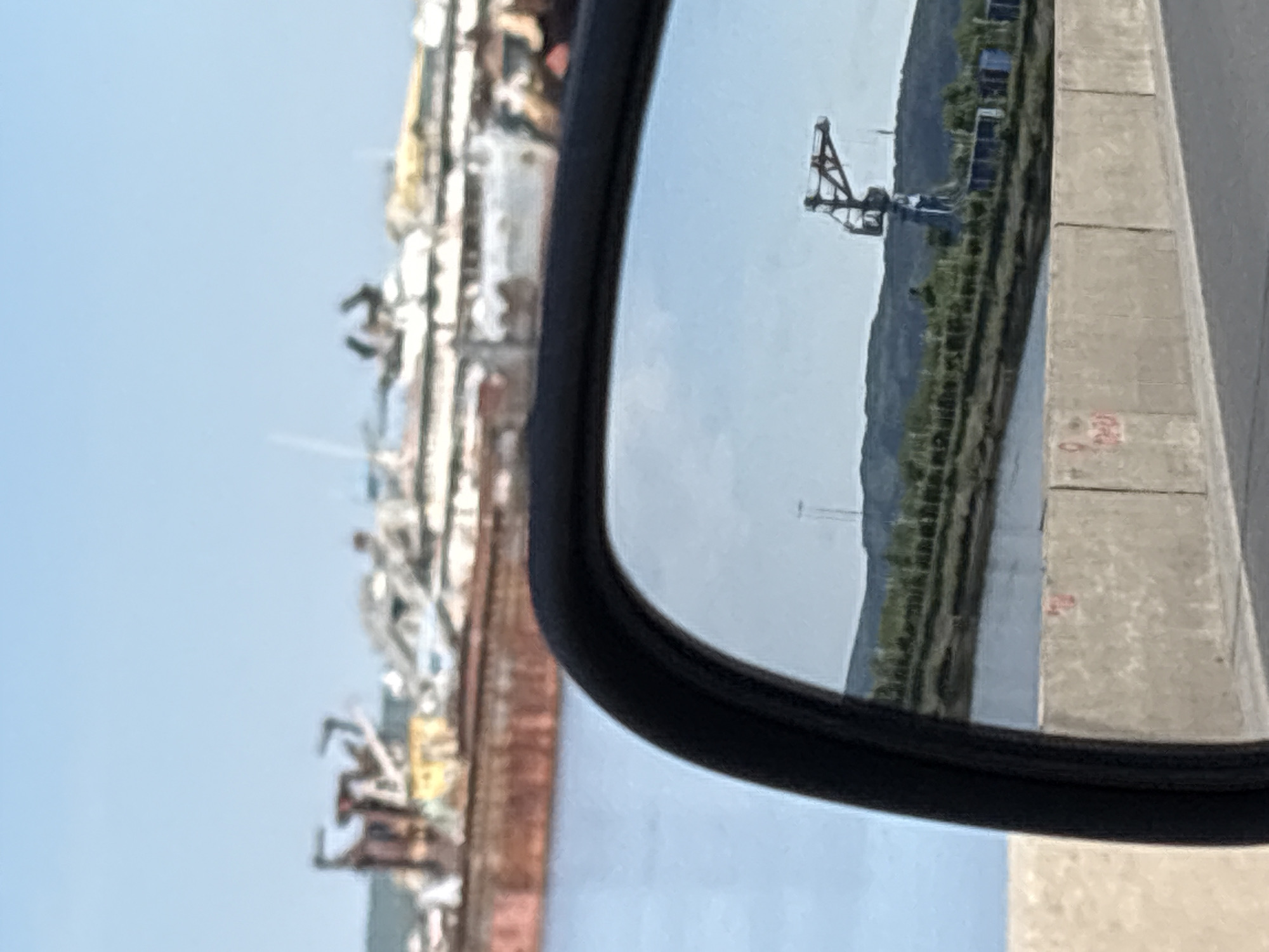 The image shows a view in a side mirror. Reflected in the mirror is a shoreline with vegetation, a waterway, concrete barriers, and an industrial crane. In the foreground, to the left of the mirror, is a blurry view of what appears to be a collection of ships or boats with some scaffolding. It is difficult to identify the exact location in Chaikovsky, Russia without more specific details or landmarks.