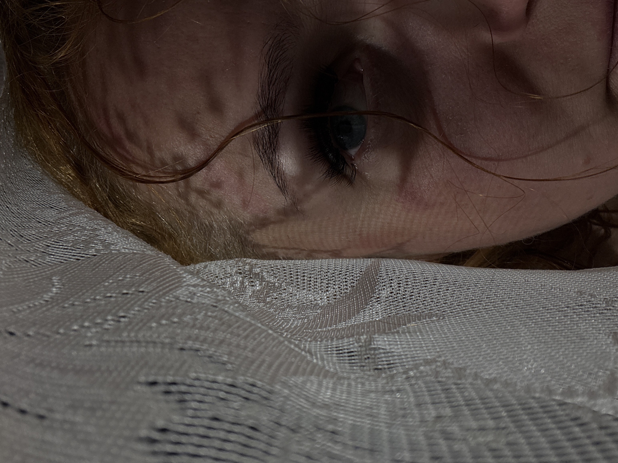 A close-up image shows a person's face partially covered by a white, textured fabric. The person's left eye is visible, displaying a blue iris. A strand of hair crosses over the eye. The skin is fair, and the overall lighting is subdued. There is no definitive evidence to place the subject or setting in Chaikovsky, Russia.