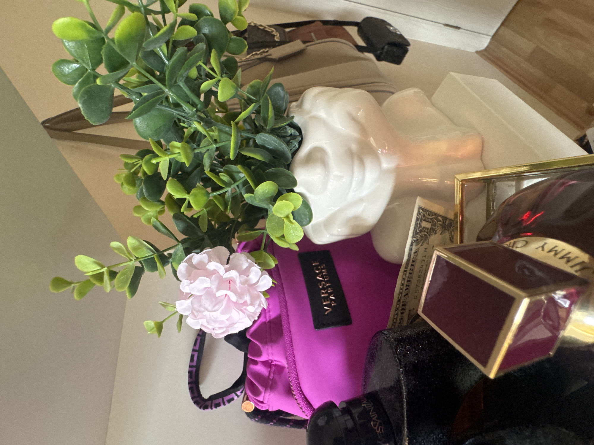 The image displays a still life composition atop a likely white surface. Dominating the scene is a pink Versace Parfums pouch, adorned with a black label bearing the brand's name. A faux green plant with a single pink flower is positioned above and to the left of the pouch. Adjacent to the Versace pouch are various objects, including a white ceramic object, a stack of US currency, and several perfume bottles including a bottle labeled Jimmy Choo.
The location is undetermined, as there is no definitive information in the image to determine the city of Sofia, Bulgaria as the location.