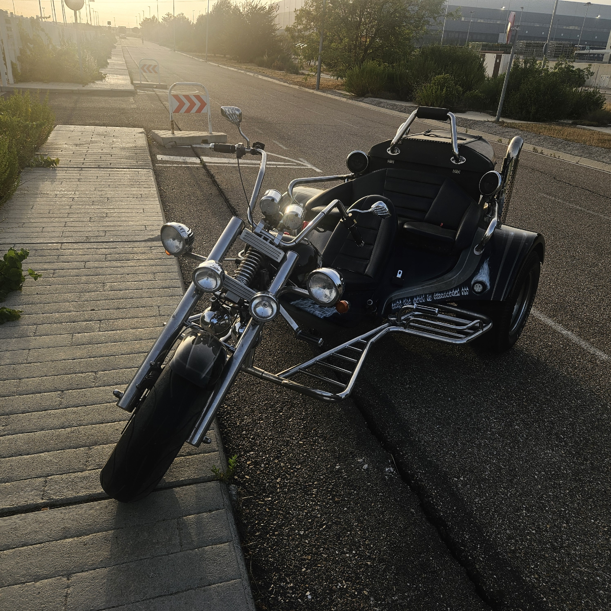 A black three-wheeled motorcycle is parked on an asphalt road in Illescas, Spain, near a paved walkway. The motorcycle has a large windshield, chrome accents, and two black seats. The sun is low in the sky, casting long shadows and creating a hazy atmosphere. In the background, there are trees, streetlights, and buildings. A few traffic signs with red and white chevron arrows are visible along the roadside. The scene appears to be a quiet street, possibly at dawn or dusk.