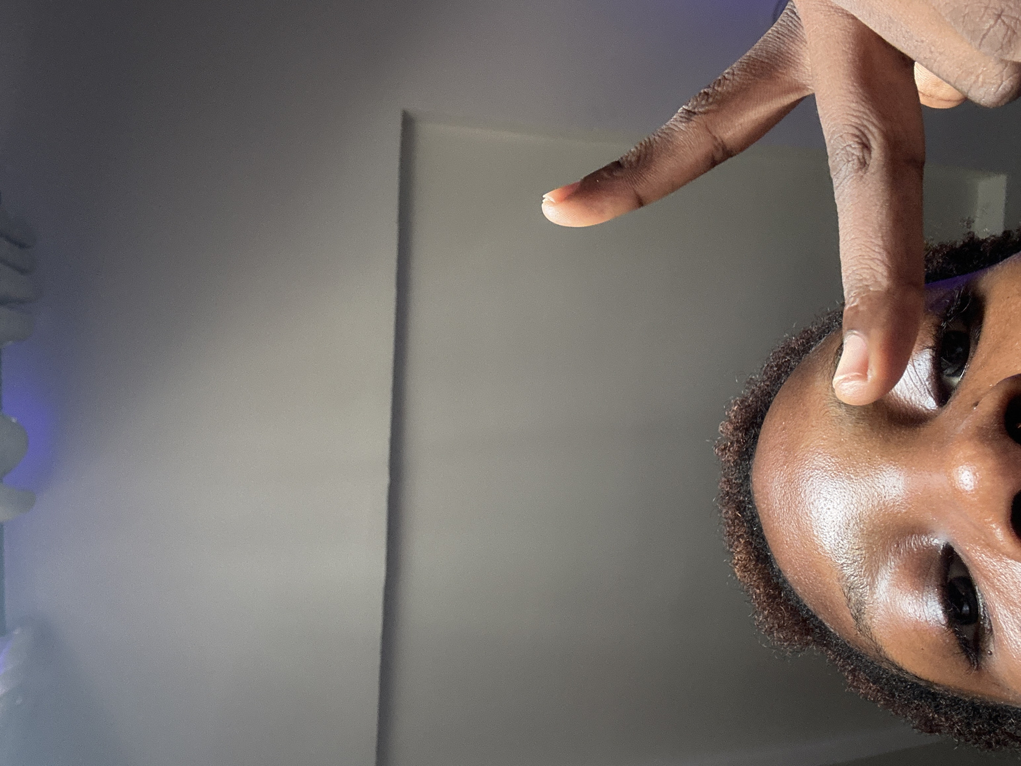 An image shows a close-up of an individual's face, with their hand raised in front of them, forming a V sign with their fingers. The skin tone of the person is dark. The image orientation is such that the person's face appears in the lower right corner, and the background is a light gray wall. There are no clear indications of a specific location in Abuja, Nigeria, or any other identifying features about the environment.