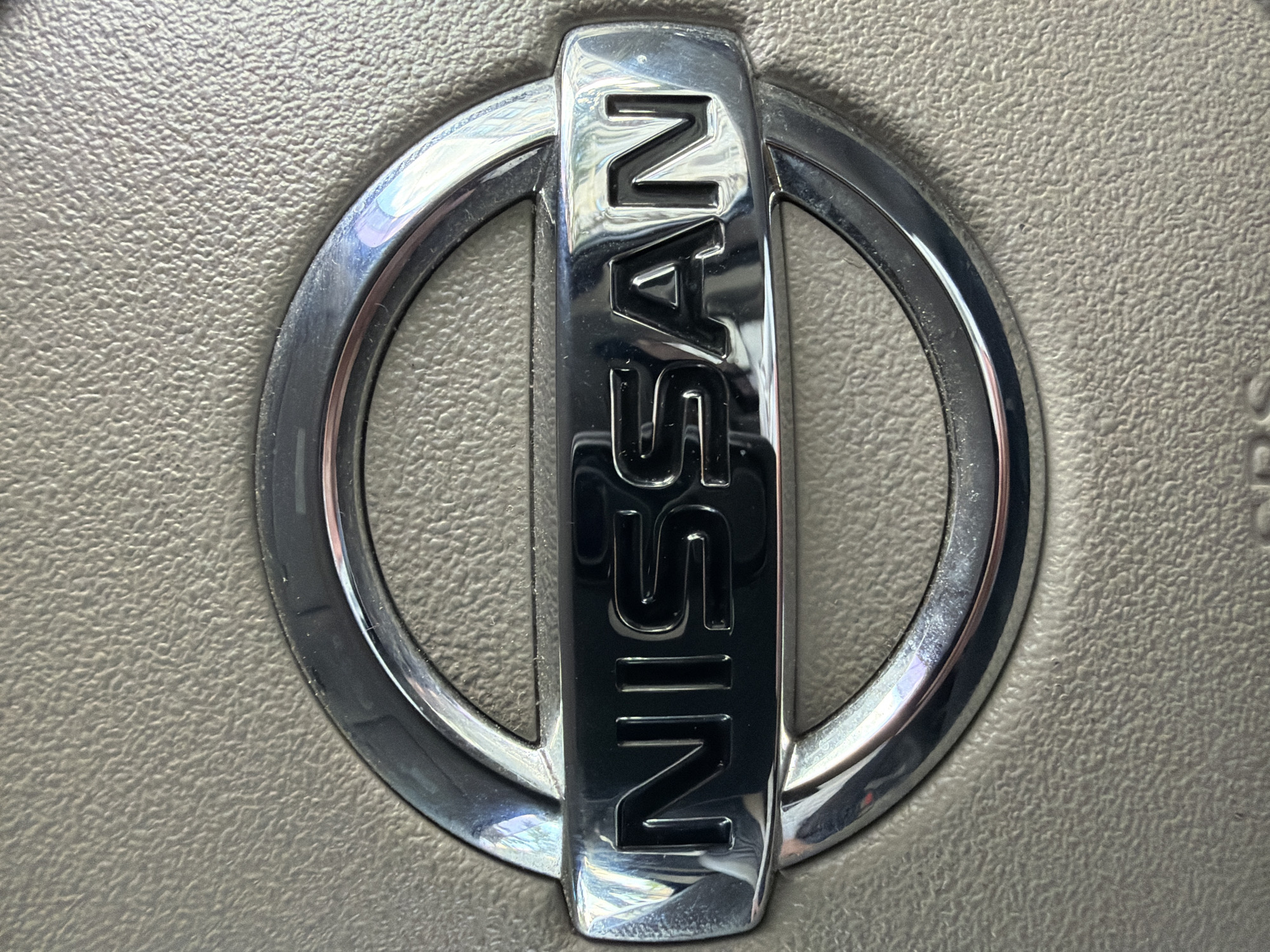 A close-up, eye-level shot features a chrome Nissan logo with the word "NISSAN" embossed in black letters on a textured, light brown surface. The logo is circular with a horizontal bar across the center, and the chrome reflects light, creating bright highlights. The textured surface appears to be part of a car's interior, possibly a dashboard or steering wheel.