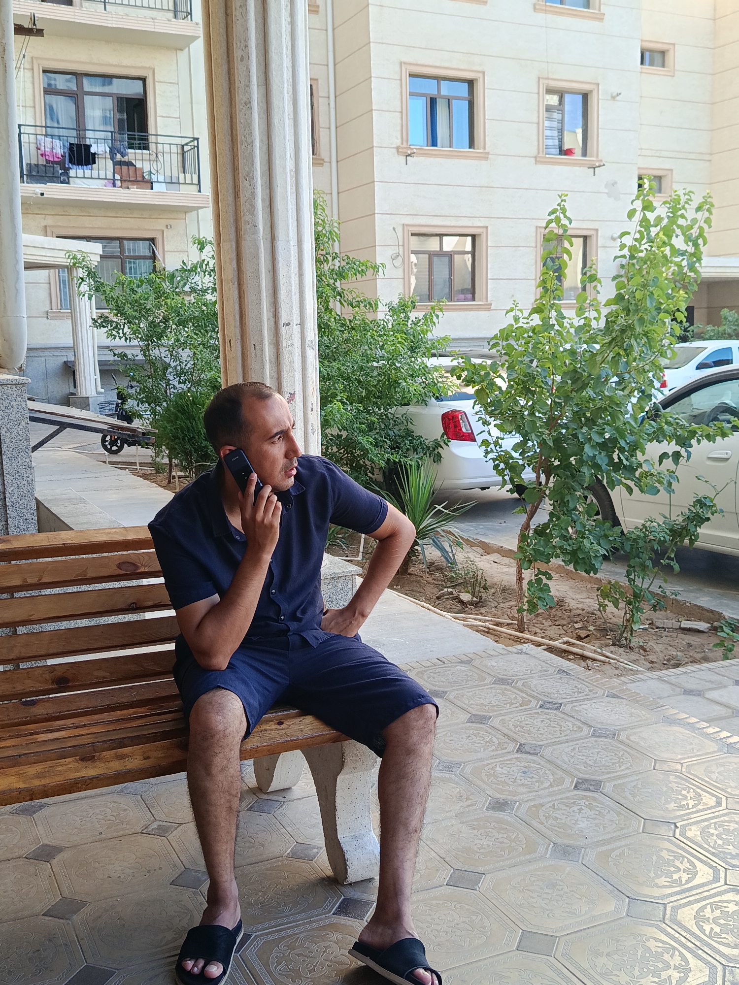 A man is sitting on a wooden bench outside a modern apartment building in Zarafshan, Uzbekistan. He is wearing a dark blue short-sleeved shirt and dark blue shorts, and black slip-on sandals. He is holding a black smartphone to his left ear and appears to be engaged in a conversation. His right hand is resting on his hip, and his left arm is bent at the elbow. The bench is positioned on a tiled patio with a decorative pattern. In the background, the light-colored facade of the apartment building has multiple windows and balconies, some with laundry hanging out to dry. Green bushes and a small tree are planted in front of the building. Two white cars are parked in the background. The scene suggests a casual, everyday moment outdoors.