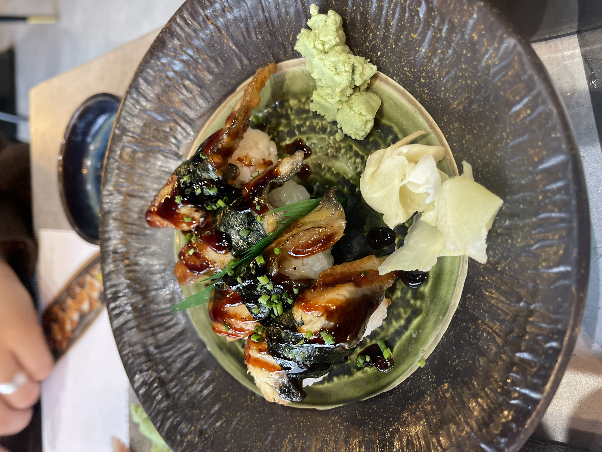 A high-angle, close-up shot shows a plate of food, likely sushi, on a table. The plate is dark, textured, with a green center holding the food. There are pieces of fish, possibly eel, drizzled with a dark sauce, placed on small rice beds wrapped with seaweed. Green onions are sprinkled on top. The plate also contains wasabi and pickled ginger. A small bowl is visible on the side. The environment suggests a restaurant setting, possibly in London, United Kingdom, given the context. A hand wearing a ring is partially visible on the left. The lighting appears to be natural.