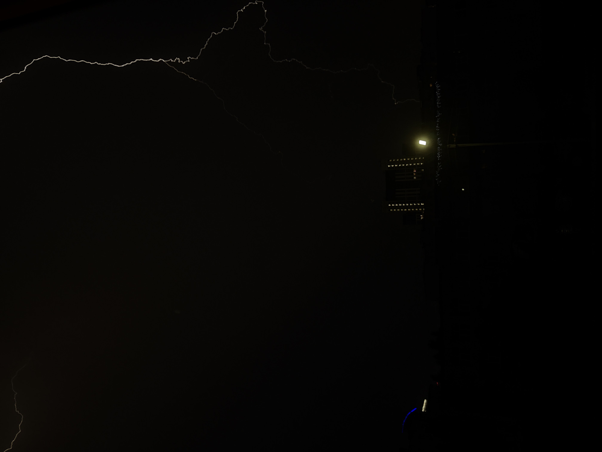 The image captures a stormy night in an unspecified urban location. A bright, jagged bolt of lightning illuminates the dark sky, arcing from the upper right to the mid-left of the frame. Below the lightning, silhouetted against the night, is a low-rise building or a cluster of buildings with illuminated windows, suggesting it's nighttime. In the foreground, a street lamp with a bright, glowing bulb stands on a pole, casting light onto the ground. The overall scene depicts a dramatic natural phenomenon occurring over a city.