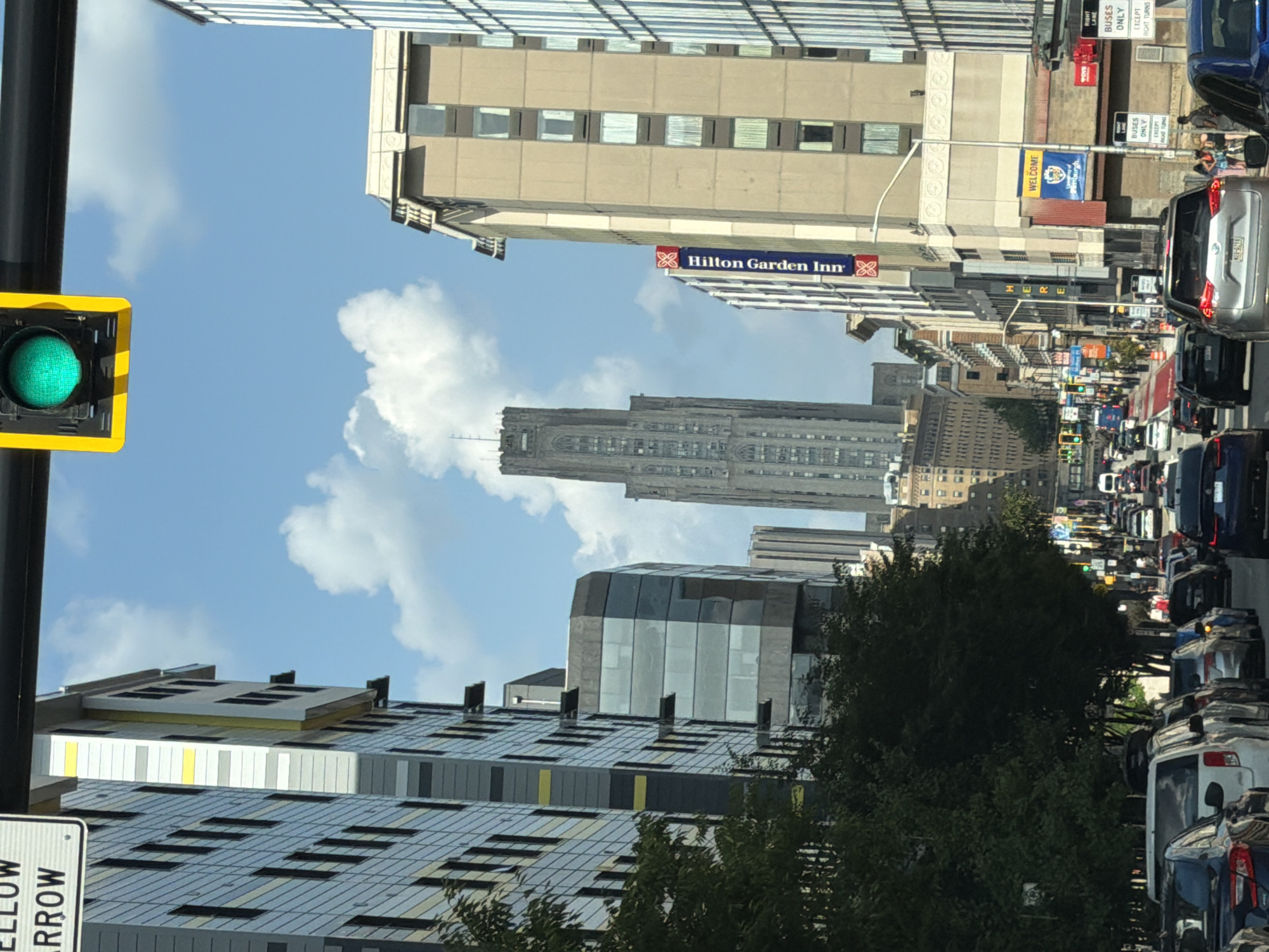The image shows a city street scene in Pittsburgh, Pennsylvania, United States of America, looking down a busy street with tall buildings on either side. A green traffic light is visible at the top of the frame. The buildings include modern high-rises with glass facades and older structures. One building prominently displays a "Hilton Garden Inn" sign. The street is filled with cars in traffic, suggesting it is a weekday or during commuting hours. Trees line sections of the sidewalk, providing some greenery amidst the urban environment. Several street signs are visible, including traffic regulations and directional information. People are seen walking on the sidewalks in the distance. The sky is blue with scattered clouds.