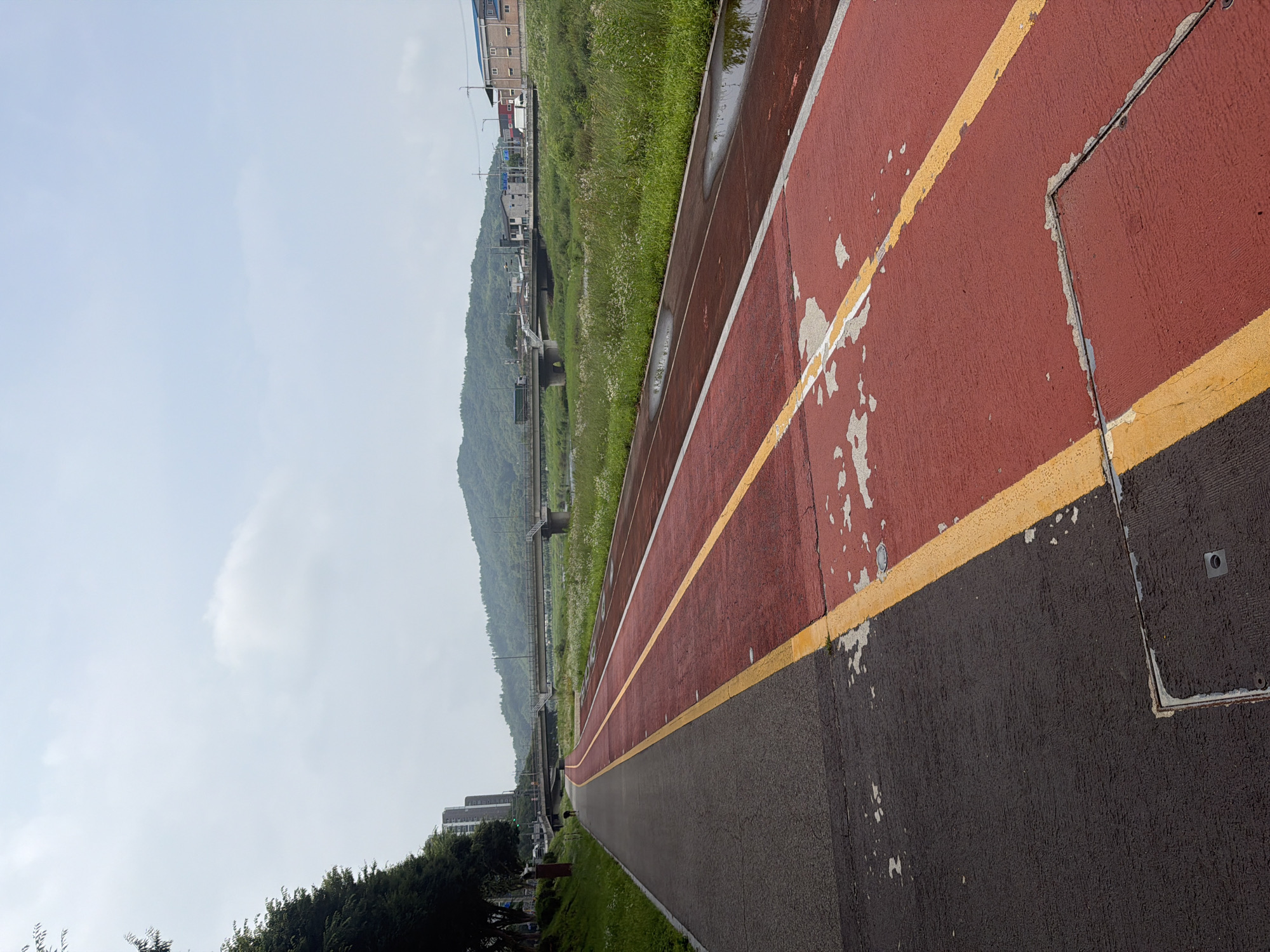 This is a photograph of a paved path next to a body of water, likely a river, in Dongducheon-si, South Korea. The path is divided into two lanes: a darker, grey lane on the left, and a red lane with yellow markings on the right. The red lane appears to be a designated sports track, possibly for running or cycling, and shows signs of wear with patches of the red surface peeling away. Several puddles of water are visible on the red lane, indicating recent rain.
Beyond the path and water, a grassy bank is lined with reeds and wildflowers. A bridge crosses the water in the background, with traffic visible on it, suggesting it is a road bridge. Further in the distance, rolling green hills form the horizon, with a few buildings and trees scattered across the landscape. The sky is overcast with diffused light. On the left side of the path, a line of trees and some taller buildings are visible, including what appears to be an apartment complex. A solitary figure, seen from behind and appearing to be walking on the grey lane, is present in the mid-left of the frame, adding a sense of scale and human presence to the scene.