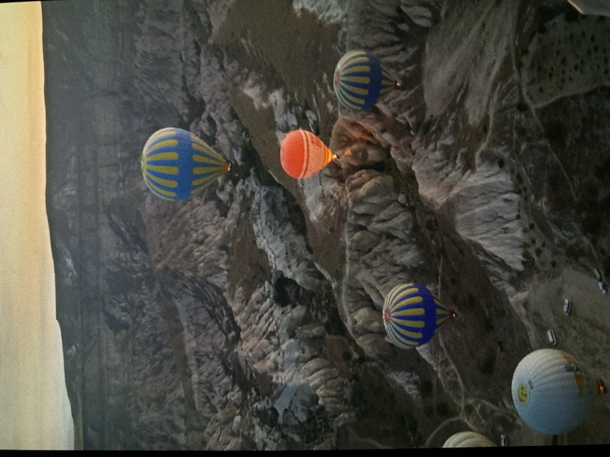 The image provides an aerial perspective of a hot air balloon flight over a rugged, eroded landscape, contextually set in Bankya, Bulgaria. Several hot air balloons are visible at various distances and altitudes. In the foreground and mid-ground, two prominent balloons feature distinctive blue and yellow vertical stripes. A bright orange balloon is positioned centrally, glowing subtly, suggesting the operation of its burner. Towards the bottom right, a white or light blue balloon is partly visible, adorned with a clear yellow smiley face icon. Another blue and yellow striped balloon appears further in the distance.
The activity depicted is hot air ballooning, showcasing multiple balloons in flight. The landscape below is characterized by complex geological formations, including steep, layered rock structures, deep valleys, and natural erosions, giving the terrain a textured and undulating appearance. Sparse vegetation or barren rock covers the ground. In the lower right corner, several vehicles, possibly cars or vans, are scattered on what appears to be a road or open area, suggesting a launch or landing site, or a spectator viewpoint.
The time of day appears to be early morning or late afternoon, indicated by the warm, soft light originating from the left side of the frame, likely sunrise or sunset. The atmospheric conditions seem clear and calm, ideal for ballooning. No notable weather events are present. The only visible text or recognizable symbol is the yellow smiley face on the white balloon. The scene conveys a sense of peaceful adventure and panoramic exploration.