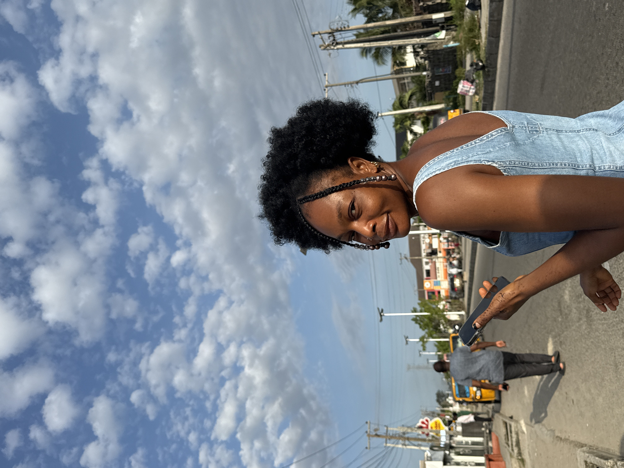 The image is taken outdoors in Ado, Nigeria, featuring a young woman in a light blue denim dress, adorned with braids and beads in her hair. She is smiling and holding a phone. In the background, there's a street with buildings, palm trees, power lines, and other vehicles including a yellow tricycle (keke). A man wearing a grey shirt and dark pants is walking in the same direction as the tricycle. The sky is blue with many white clouds.