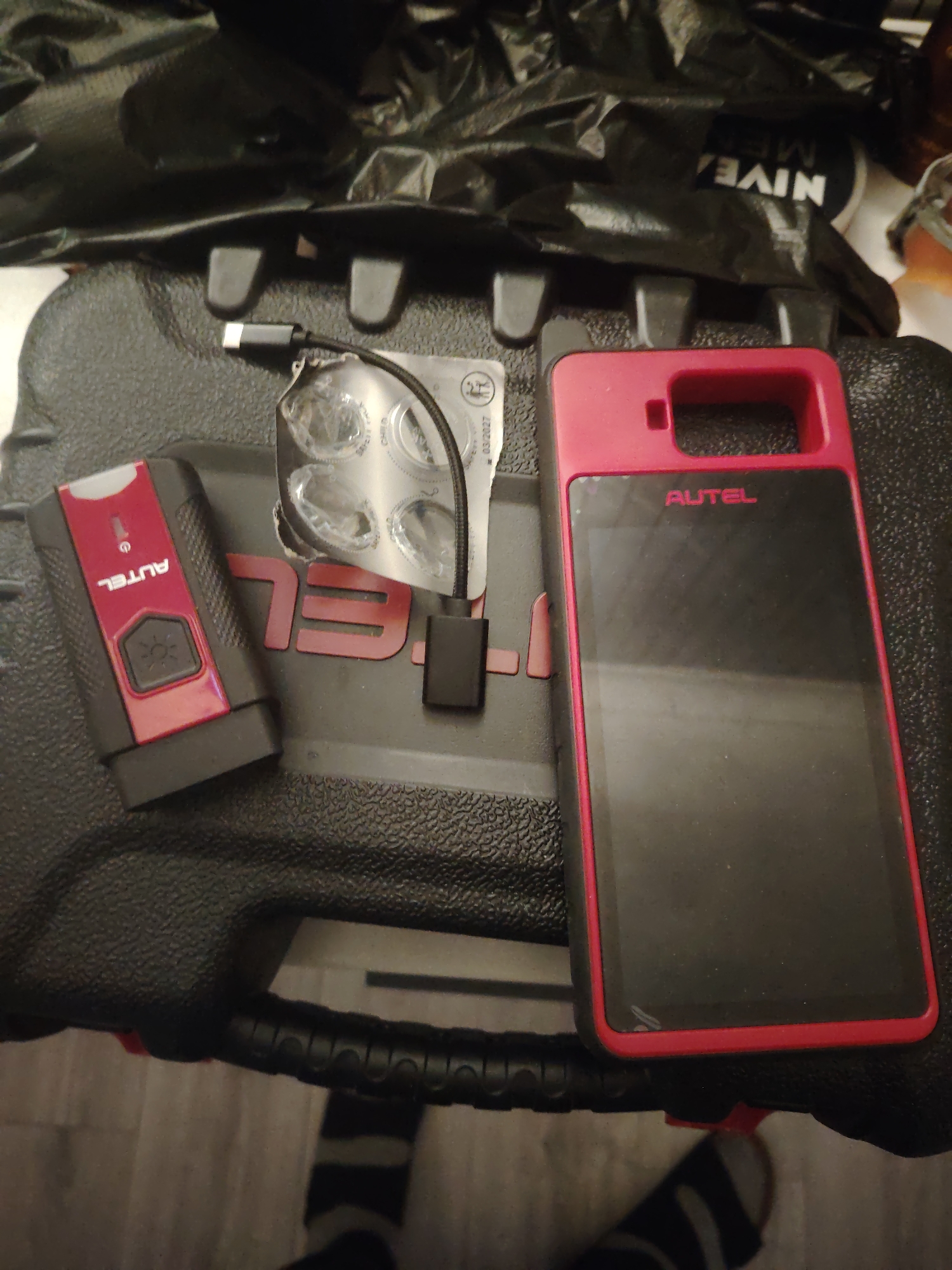 An overhead shot captures various electronic devices and packaging materials laid out on a dark, textured surface, possibly a protective case, in an indoor setting in Huntingdonshire, United Kingdom.
The primary subjects are two black and red electronic devices from the brand AUTEL. On the left is a smaller, rectangular device, likely an automotive diagnostic scanner, with "AUTEL" printed vertically in white on a red section. It features a black button with a white sun-like symbol. To its right lies a larger, tablet-like device or smartphone enclosed in a robust red and black case, also displaying "AUTEL" in red near the top of its black screen.
Between these two devices are a coiled USB-C cable and a small black USB-A adapter. Adjacent to them is a metallic blister pack, possibly containing batteries or small components, with the date "03/2027" and partially visible text "CHILD RESISTANT PACKAGING". Behind these items, a crumpled black plastic bag is visible, bearing the reversed white text "NIVEA MEN".
The items appear to be either in preparation for use, in the process of being packed away, or recently unpacked. A foot wearing a black and grey striped sock is partially visible in the bottom right corner, suggesting human presence nearby. The overall lighting suggests an interior space, with no specific time of day or weather conditions discernible from the image.
