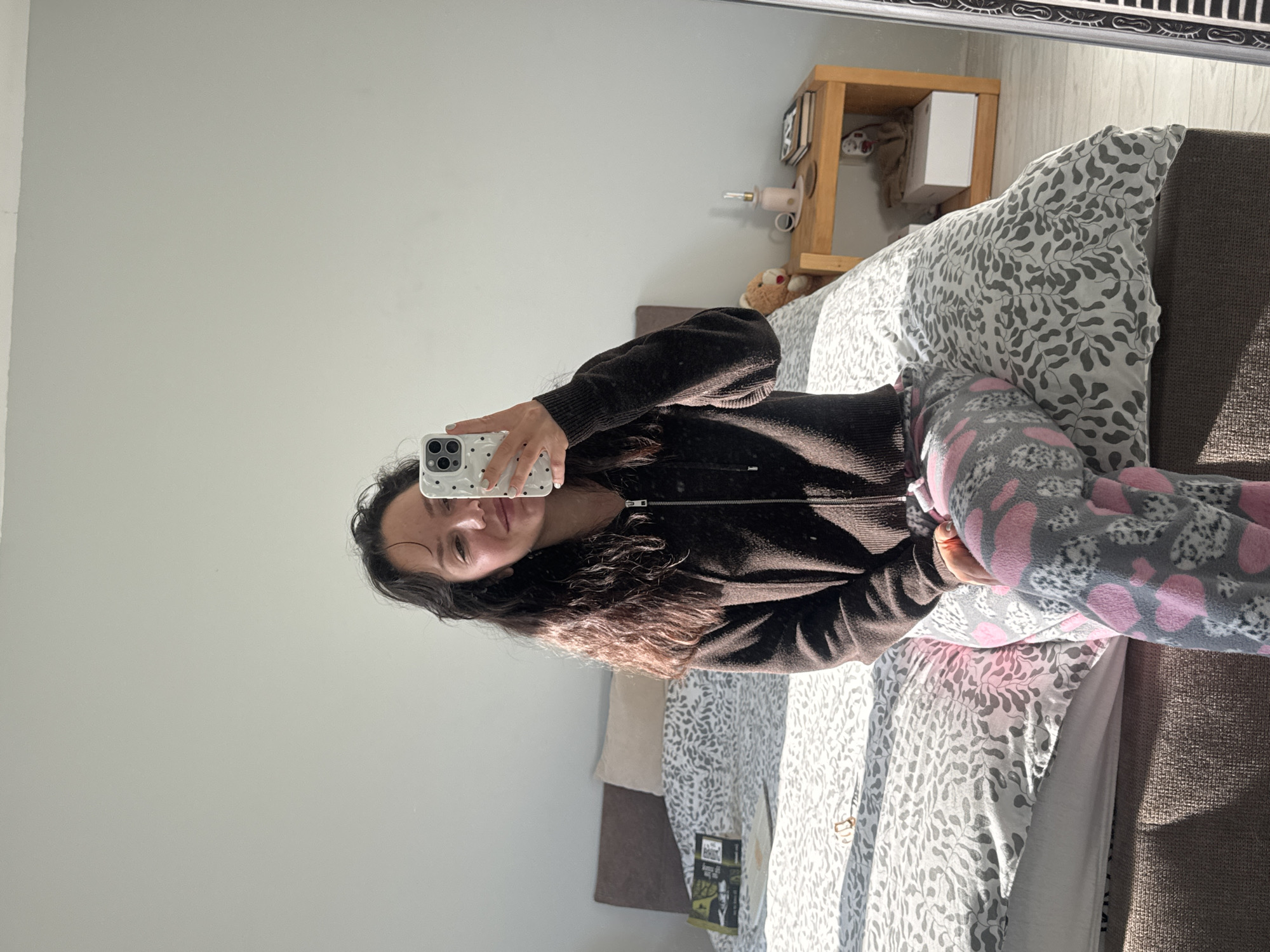 A young woman is seen taking a mirror selfie in a brightly lit bedroom in Burgas, Bulgaria. She is lying or sitting on a bed, holding a white smartphone with black polka dots, angled to capture her reflection. Her long, dark, wavy hair frames her face as she looks towards the phone. She is dressed in a dark brown or black zippered top and comfortable grey and pink patterned pajama bottoms, possibly featuring hearts or animal shapes. Her fingernails are painted a light greenish-blue.
The room is well-lit by natural daylight, casting soft shadows, indicating a sunny day. The bed is adorned with a duvet cover featuring a grey leafy pattern on a white background. To the right of the bed, a light brown wooden nightstand or shelf is visible, holding a small white box, a teddy bear, and a pink-colored object, possibly a small lamp or diffuser. The walls are a light, neutral color. A book or magazine, partially visible on the bed, has some text on its cover, but it is not clear enough to read. The overall scene suggests a relaxed and casual moment in a personal living space.