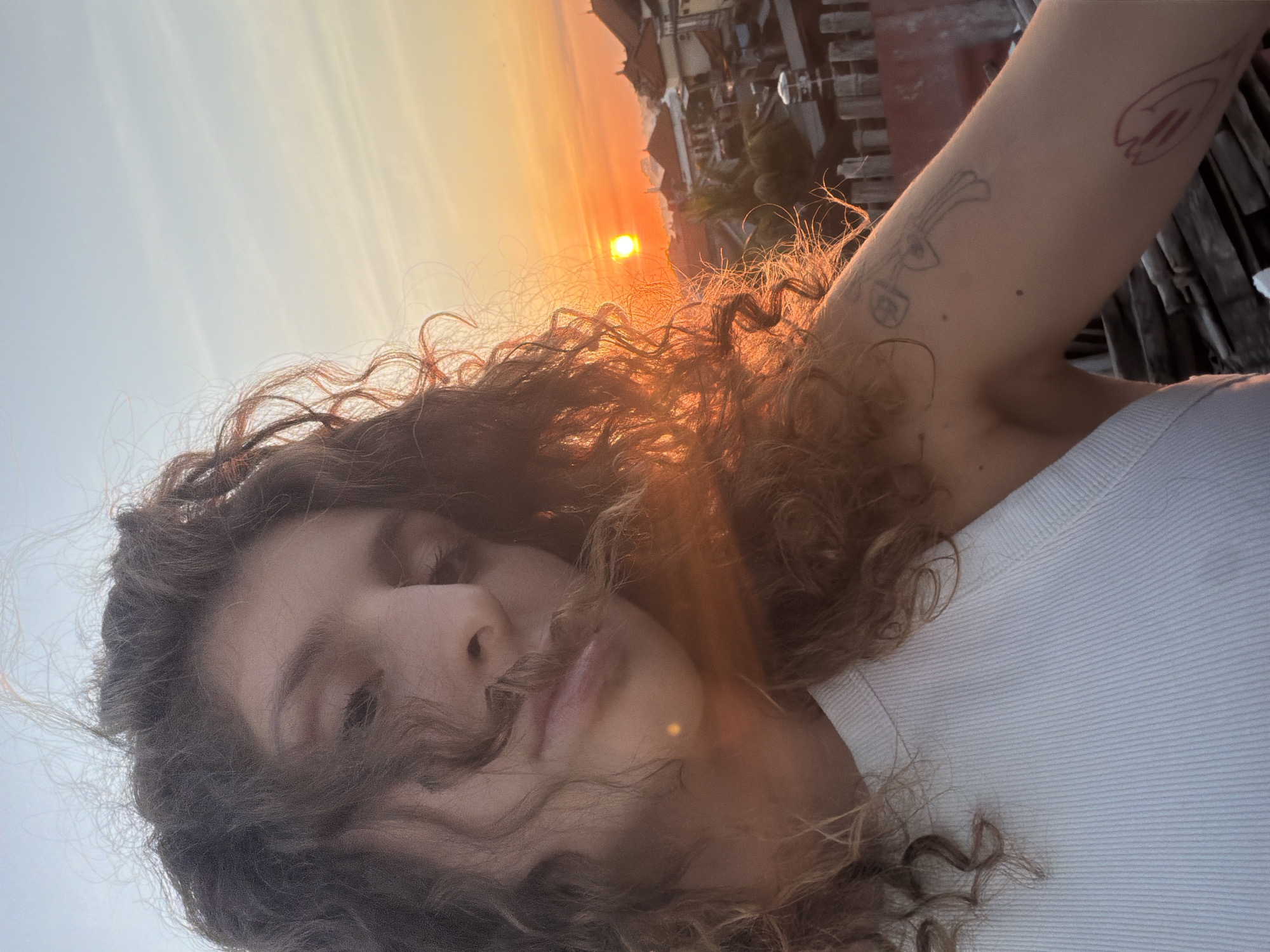 A selfie of a woman with curly hair is taken outdoors against a backdrop of a sunset. Her upper body is visible, showing a white ribbed tank top and a fair-skinned arm with two distinct tattoos. The background includes silhouettes of buildings, and the sun is prominently featured in the upper center. The picture is taken in North Kuta, Indonesia.