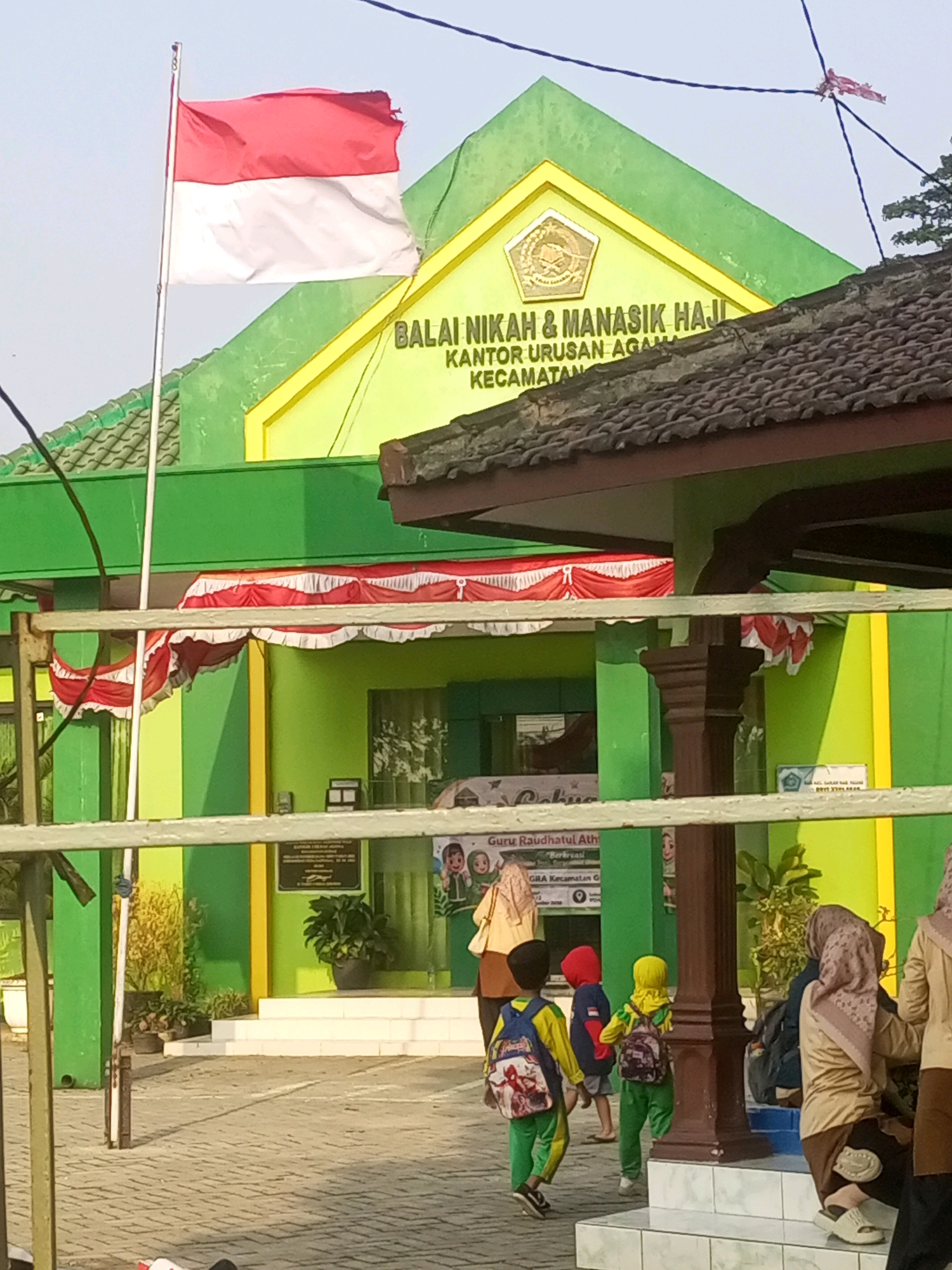 In the city of Gurah, Indonesia, a building with green and yellow accents is visible. The building has a prominent sign reading "BALAI NIKAH & MANASIK HAJI KANTOR URUSAN AGAMA KECAMATAN." In front of the building, an Indonesian flag flies on a flagpole. A group of people, including several children wearing backpacks, are walking towards the entrance of the building. The children are dressed in colorful clothing, and some are wearing headscarves. The building appears to be a government office or community center. The overall scene suggests a typical day at a public facility in Indonesia, with people going about their daily activities.
