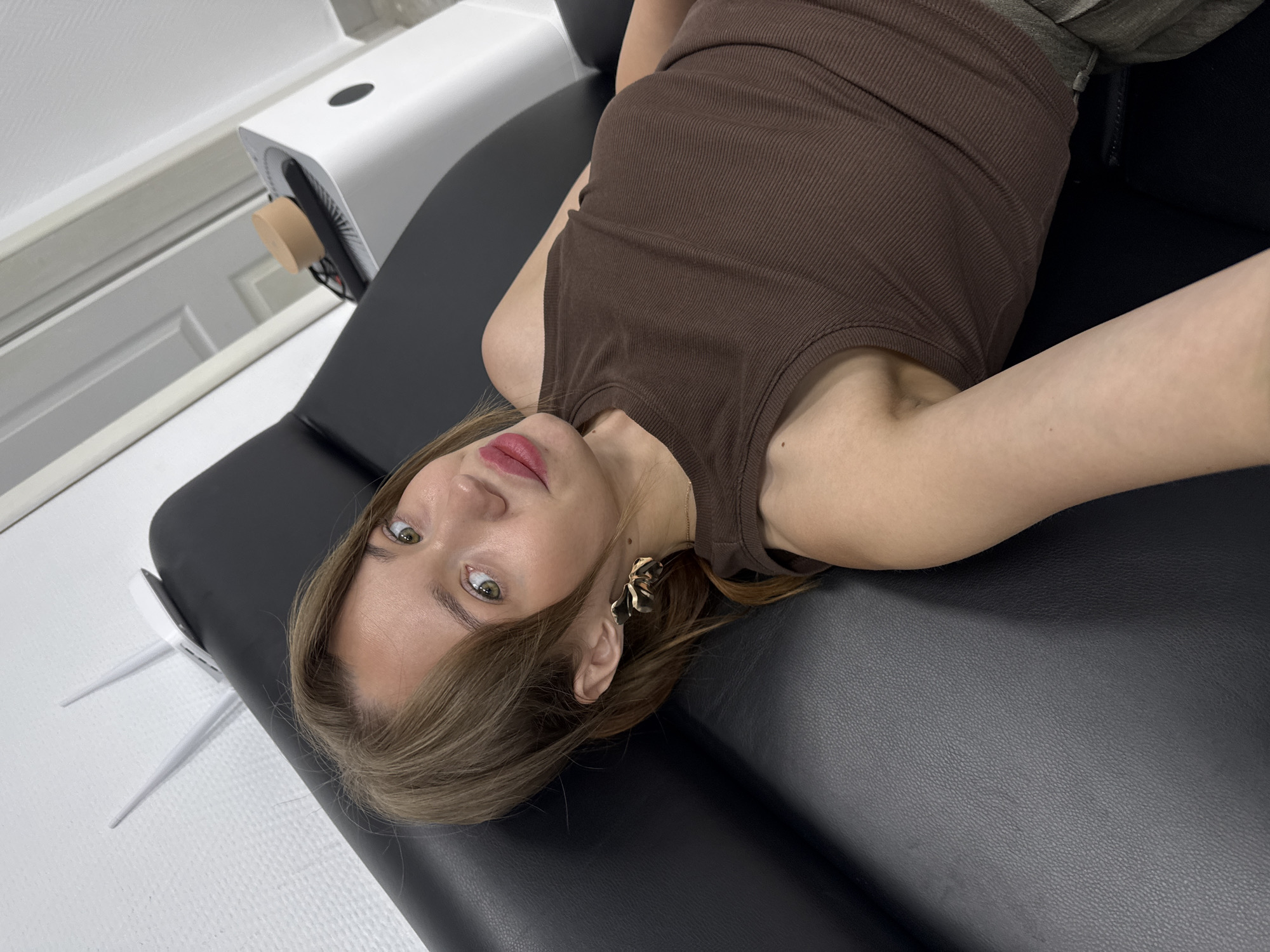 In Chaikovsky, Russia, a young woman with light brown hair and green eyes is lying on a black leather sofa. She is wearing a brown ribbed tank top and olive green pants. Her hair is pulled back into a ponytail, and she is wearing large gold earrings. Her lips are painted with pink lipstick, and she is looking towards the camera. Behind her, on the wall, is a white Wi-Fi router with two antennas. To the right of the sofa is a white air purifier with a wooden top. The lighting in the room is bright, suggesting it is daytime.