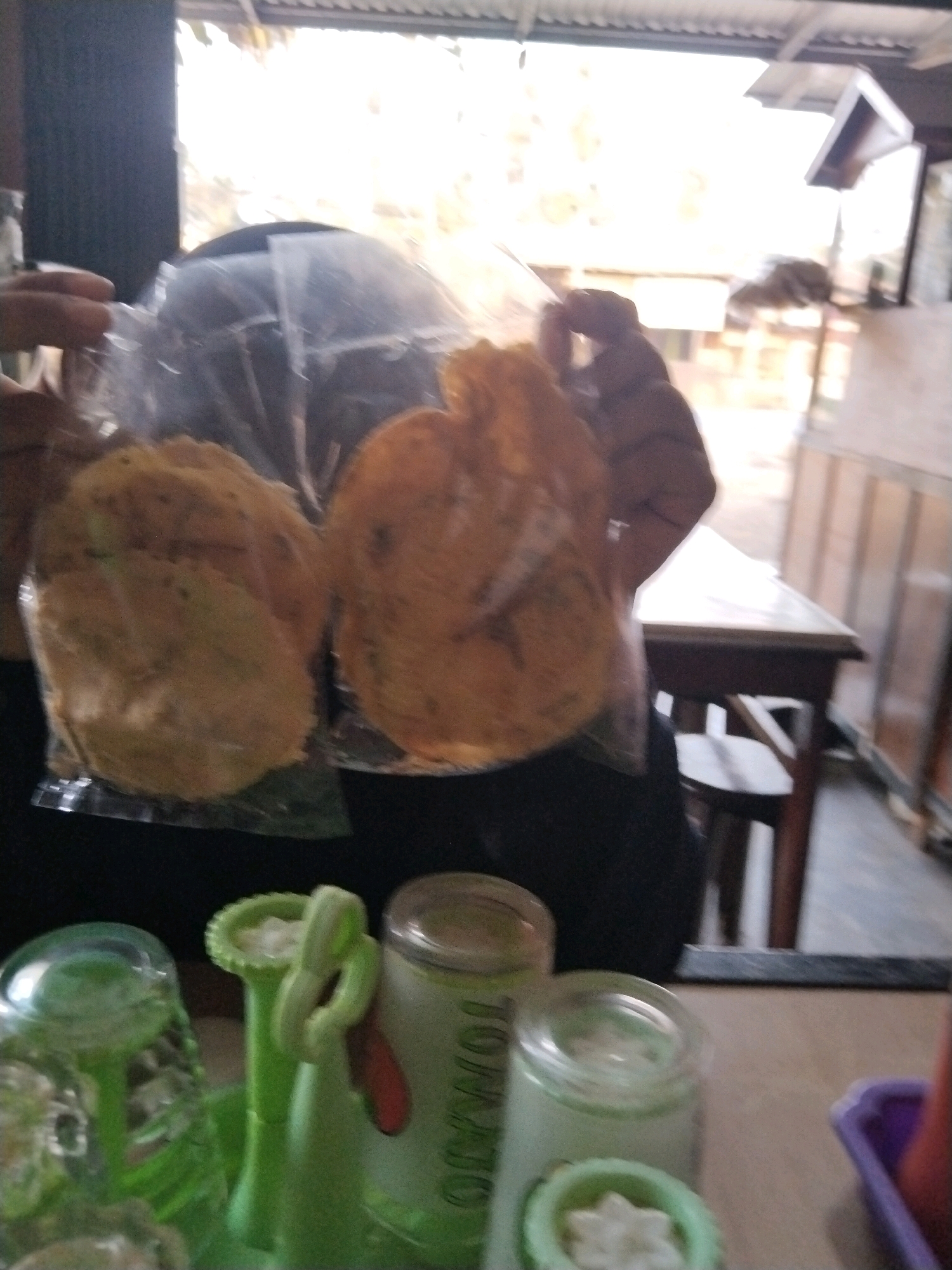 In an indoor setting that appears to be a food stall or eatery in Unknown, Indonesia, a person's hands are holding two clear plastic bags containing what appear to be fried crackers or chips. The crackers are circular and golden brown. In the foreground, several containers are visible. One is a clear plastic glass with a green plastic holder. Next to it is a green, decorative item with a looped handle. Further right, a glass jar with a metal lid contains a white substance and has green lettering that includes "TOMATO" partially visible. Another glass jar with a metal lid is to the right of the "TOMATO" jar, and a purple plastic container is on the far right. In the background, a wooden table and chairs are visible, along with an open window or doorway showing daylight and a corrugated metal roof. The lighting suggests it is daytime.