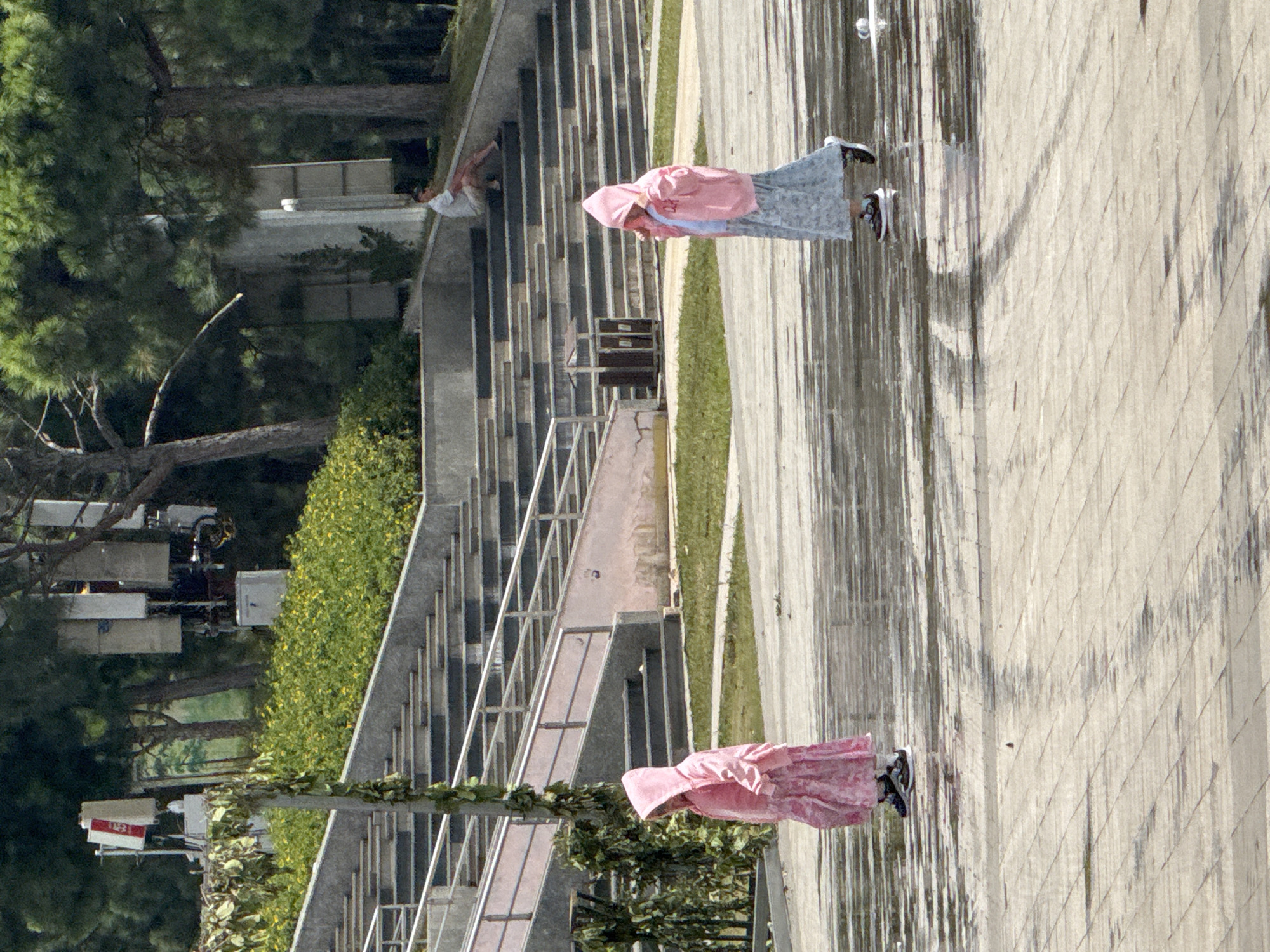 Two young girls, one on the left and one on the right, are walking on a wet paved area. The girl on the left is wearing a pink hooded jacket and a pink patterned skirt. The girl on the right is wearing a pink hooded jacket and a light blue patterned skirt. Both girls have dark shoes. They are walking away from the viewer in an outdoor setting. In the background, there are tiered concrete stairs, some with metal railings, leading up to an area with trees and bushes. There is also a person sitting on the stairs in the background, wearing a white shirt and shorts. In the far background, there are several large communication antennas mounted on poles, and lush green trees. The ground appears wet, reflecting the surroundings and casting shadows. This scene is located in Da Lat, Vietnam, likely within a park or public space with recreational facilities.