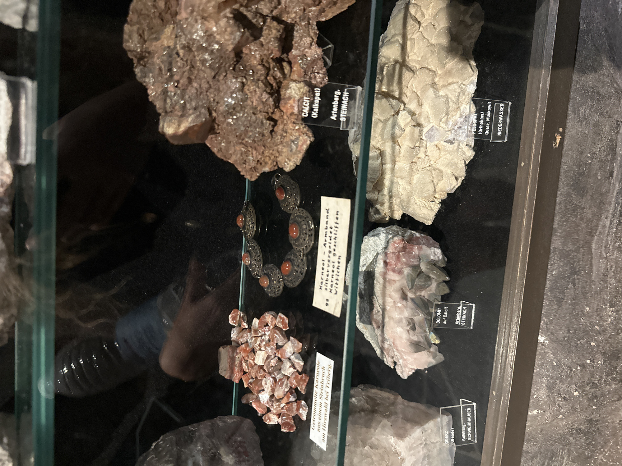 The image displays a museum exhibit of minerals behind a glass case. Several distinct rock specimens are visible, each with a small label identifying it. In the foreground, to the left, is a large, pale, crystalline rock. To the right of this is a specimen of white and pink crystals, also with a label. Further back, on the left, is a pile of small, reddish-pink, tumbled stones. Next to these is a piece of jewelry featuring multiple circular metal ornaments with reddish-orange stones. To the right of the jewelry is a large, rough, brown mineral specimen with a label. Directly below this, on the shelf, is a light-colored, crystalline mineral formation with a label. On the far right, a large, pale, layered mineral formation is visible with a label below it.
The labels on the specimens provide specific information:
- "CALCIT (Kalkspat) Artenberg, STEINACH"
- "QUARTZ (Geode) Gaisberg, SCHWIGHÄUSEN"
- "DOLOMIT auf Calcit Artenberg, STEINACH"
- "FELDSPAT (Orthoklas) Quarz, Muskovit NIEDERWASSER"
- "Getrommelte Karneole aus einem Steinbruch am Hirzwalld bei Triberg"
- "Karneol – Armband silbervergoldet 99 Karneol geschliffen Wittichen"
The reflections on the glass indicate the presence of people and lighting. A person with blonde, curly hair is visible reflected in the top portion of the glass, appearing to be standing behind the exhibit. The overall environment suggests an indoor display, likely a museum or a geological exhibition. The city of Schonach im Schwarzwald is mentioned as the location, and the labels themselves indicate specific geographical origins for some of the specimens, such as Triberg, Artenberg, Steinach, Gaisberg, Schwighausen, and Niederwasser, all locations within or near the Black Forest region of Germany.