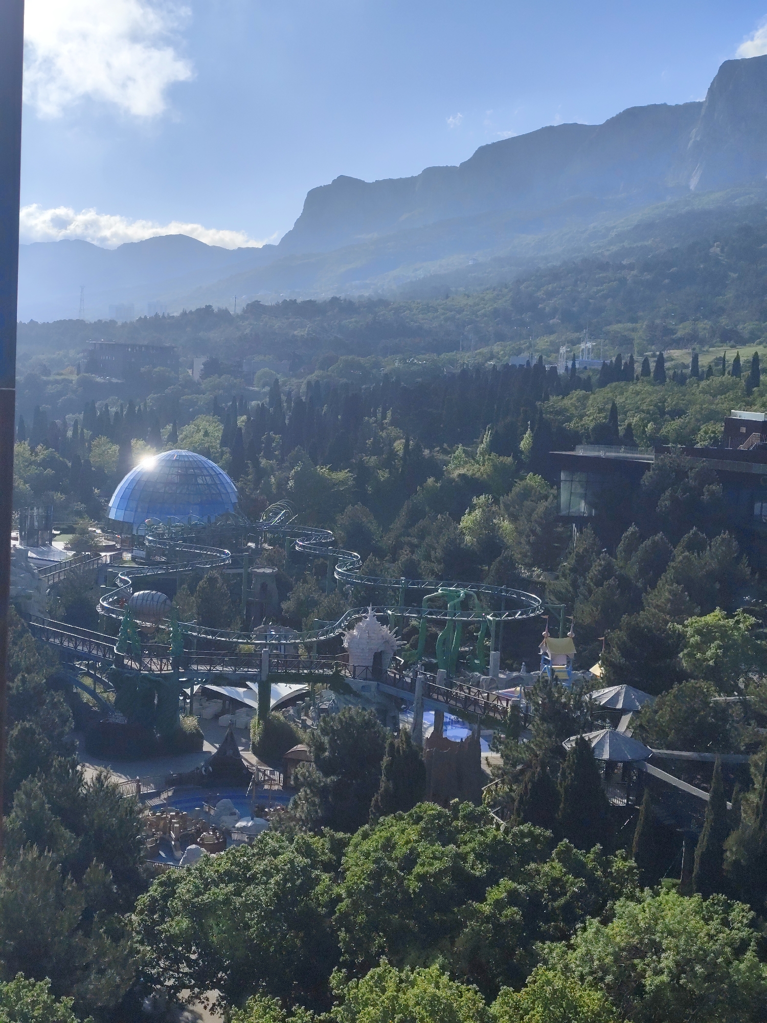 The image shows a panoramic view of a park or entertainment complex set against a backdrop of mountains. In the foreground, dense vegetation, primarily trees, fills the lower portion of the frame. Within the trees is a theme park, featuring a blue geodesic dome structure, roller coasters, and various buildings and attractions. The roller coaster tracks wind around the dome and other structures, creating a complex network of paths. The background features large mountains, with some areas covered in trees and others appearing rocky and steep. The sky is partly cloudy, with sunlight illuminating the scene. The location is in Симеизский поселковый совет, Ukraine.
