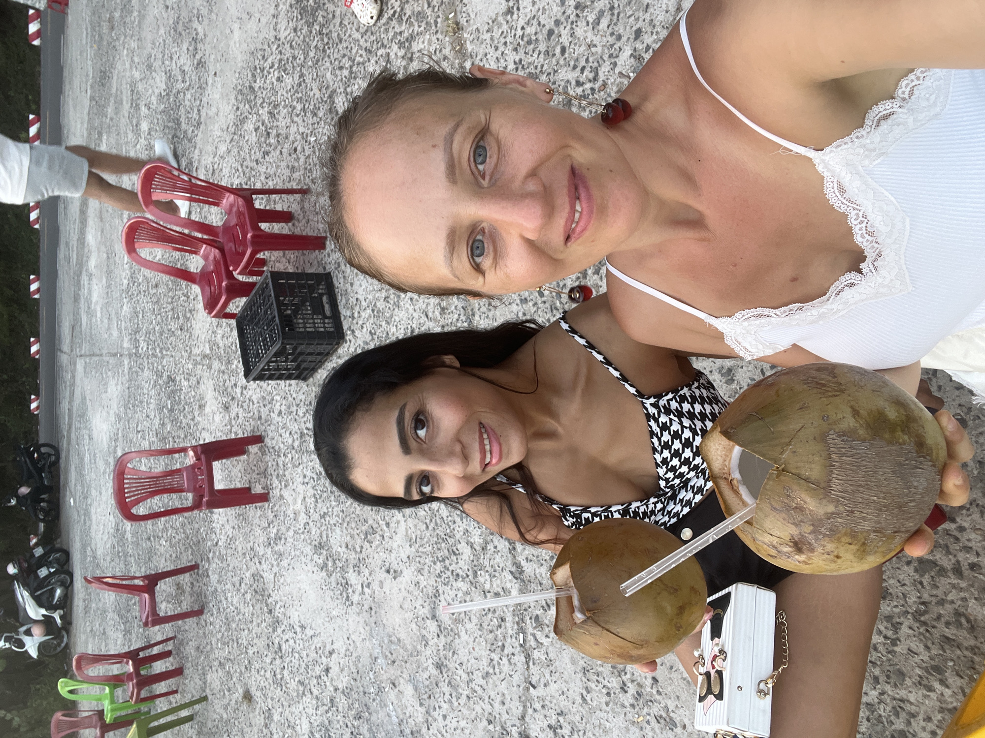 Two women are seated on the ground holding coconuts with straws. The woman on the left has long dark hair and is wearing a black and white patterned top. The woman on the right has blonde hair and is wearing a white camisole with lace trim. Both women are smiling at the camera. Behind them are several red plastic chairs and a black plastic crate. In the background, there are parked motorcycles and a road with white and red striped barriers. The ground is paved with a rough, grey material. This scene appears to be a roadside stall or rest stop, possibly along the coast of Đà Nẵng, Vietnam, given the presence of parked scooters and the tropical coconuts.