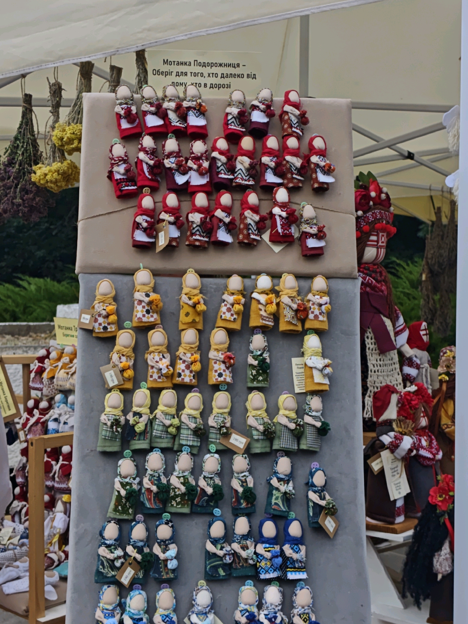The image depicts a market stall in Vinnytsia, Ukraine, selling traditional Ukrainian motanka dolls. The stall is set up outdoors, likely under a tent for shade, with the background showing greenery and other market elements. The primary focus is a large display board covered in brown paper, on which numerous motanka dolls are arranged in rows.
The dolls are handmade fabric figures, mostly small and doll-sized. They are dressed in traditional Ukrainian clothing, characterized by vibrant colors, intricate patterns, and embroidery. Many of the dolls have red, white, and patterned outfits, with some featuring red head coverings or scarves. The dolls vary in their specific designs and accessories, with some holding small objects or flowers.
Above the dolls, a sign written in Ukrainian reads: "Мотанка Подорожниця - Оберіг для того, хто далеко від дому, хто в дорозі" which translates to "Motanka Traveler - A talisman for someone far from home, for someone on the road." This indicates the cultural significance and purpose of these dolls as protective charms for travelers.
To the left of the main display, more motanka dolls are visible on a wooden shelf, some with price tags attached. In the background, bundles of dried herbs and flowers hang, typical of folk craft or herbalist stalls. To the right, a larger motanka doll, standing upright, is dressed in more elaborate traditional attire, including a detailed embroidered shirt and a headpiece. Further to the right, other market goods and stalls can be seen, suggesting a bustling outdoor market environment. The lighting suggests it is daytime.