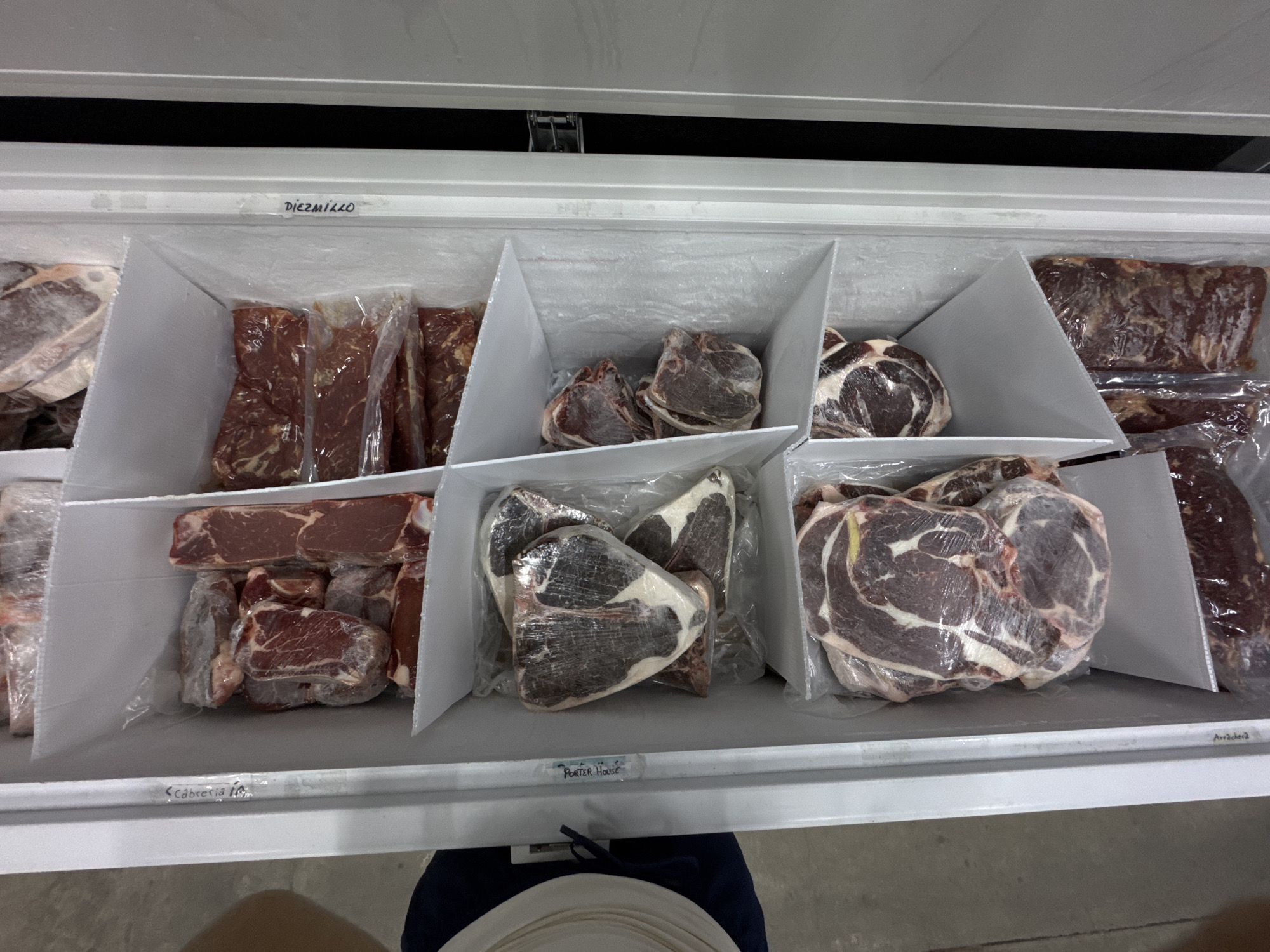 The interior of a commercial freezer is seen from an overhead perspective. The freezer is partitioned into sections with white dividers, each containing different cuts of meat, individually wrapped in clear plastic. In the front of the freezer are labels indicating the type of meat in each section: "Cabrería," "Porter House," "Diezmillo," and "Arrachera." The meats vary in appearance, some appearing leaner and more uniformly red, while others have significant marbling and fat content. The bottom of a person wearing dark-colored pants with a lighter-colored object on top, possibly plates, is visible in the lower part of the frame. There is no evidence suggesting the city of Puebla, Mexico or other city or country.