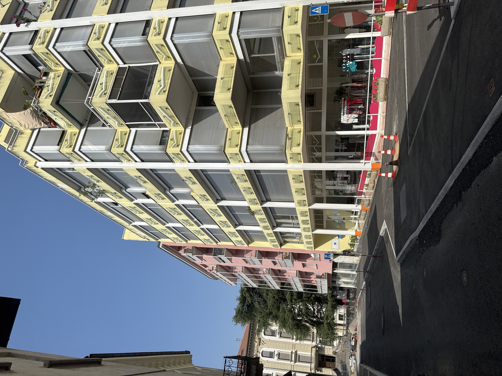 The image depicts a street scene in Chiasso, Switzerland, with modern buildings on either side and a clear blue sky overhead. The foreground shows a freshly paved asphalt road with white lane markings and some traffic cones, indicating ongoing construction or road work. To the right of the road, there is a sidewalk with planters and a store with a large glass facade displaying clothing. The building to the right is a bright yellow, multi-story structure with a distinctive facade featuring balconies with decorative yellow panels and white railings. The building to the left is a lighter colored building, followed by a pink multi-story residential building with balconies. In the background, another ornate building with classical architectural elements is visible, partially obscured by a large evergreen tree. The overall impression is a contemporary urban environment with a mix of architectural styles.