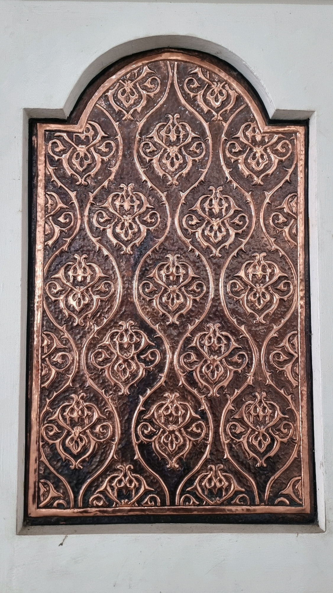 The image shows a close-up view of a decorative metal panel with an intricate, repeating arabesque pattern. The metal is a coppery-bronze color, with areas of darker patina that highlight the raised design. The pattern consists of swirling floral motifs and curved lines, forming a symmetrical and ornamental composition. The panel is framed by a dark border and appears to be set within a light-colored, possibly painted, architectural element. The background suggests an indoor setting, perhaps a wall or a doorway. The image does not contain any people or indicate any specific actions or interactions. The location is not discernible from the image itself, and therefore cannot be specified for Lhokseumawe, Indonesia.