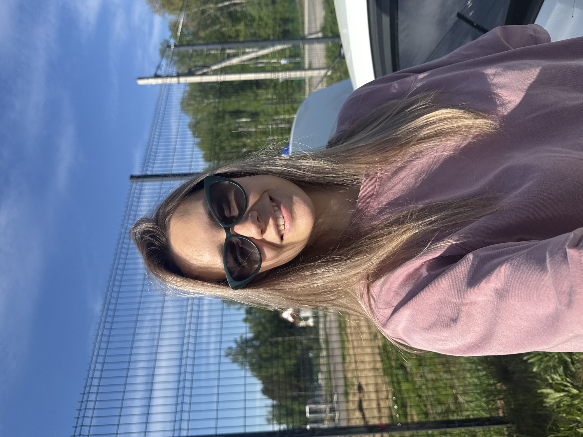 A woman with long, blonde hair and sunglasses is smiling at the camera. She is wearing a pink sweatshirt. A wire fence and greenery can be seen in the background. The location appears to be Noviy, Russia.