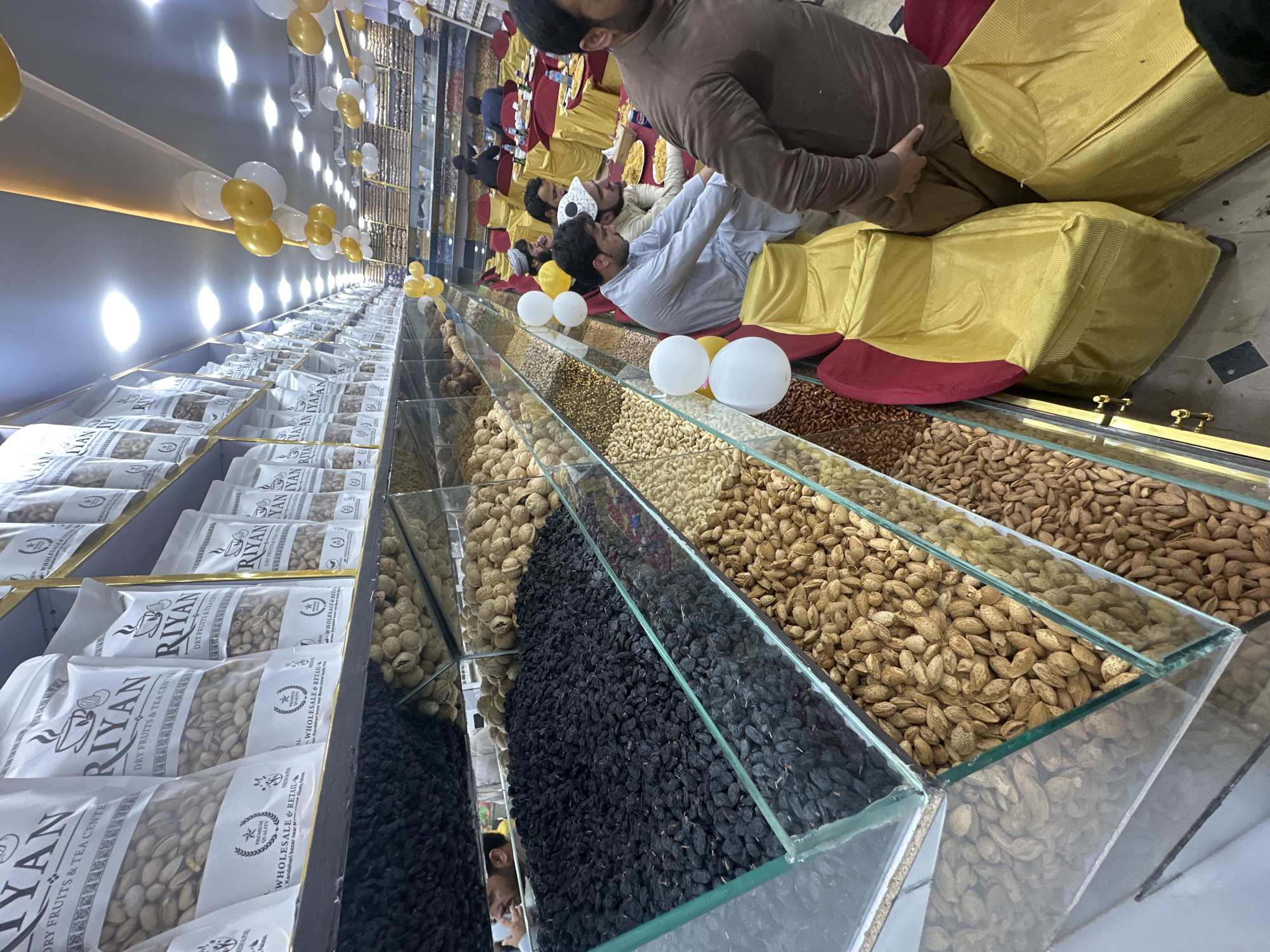 The image shows the interior of a dry fruit and tea shop in Quetta City Tehsil, Pakistan. A long display case filled with nuts, dried fruits, and tea is showcased. The products are arranged in rows and columns within glass containers. Labels are visible on the packaged products, with "Riyan Dry Fruits & Tea Center" prominently displayed. A few men are sitting on golden yellow cushions along the display case. Balloons in gold and white colors are used as interior decorations.