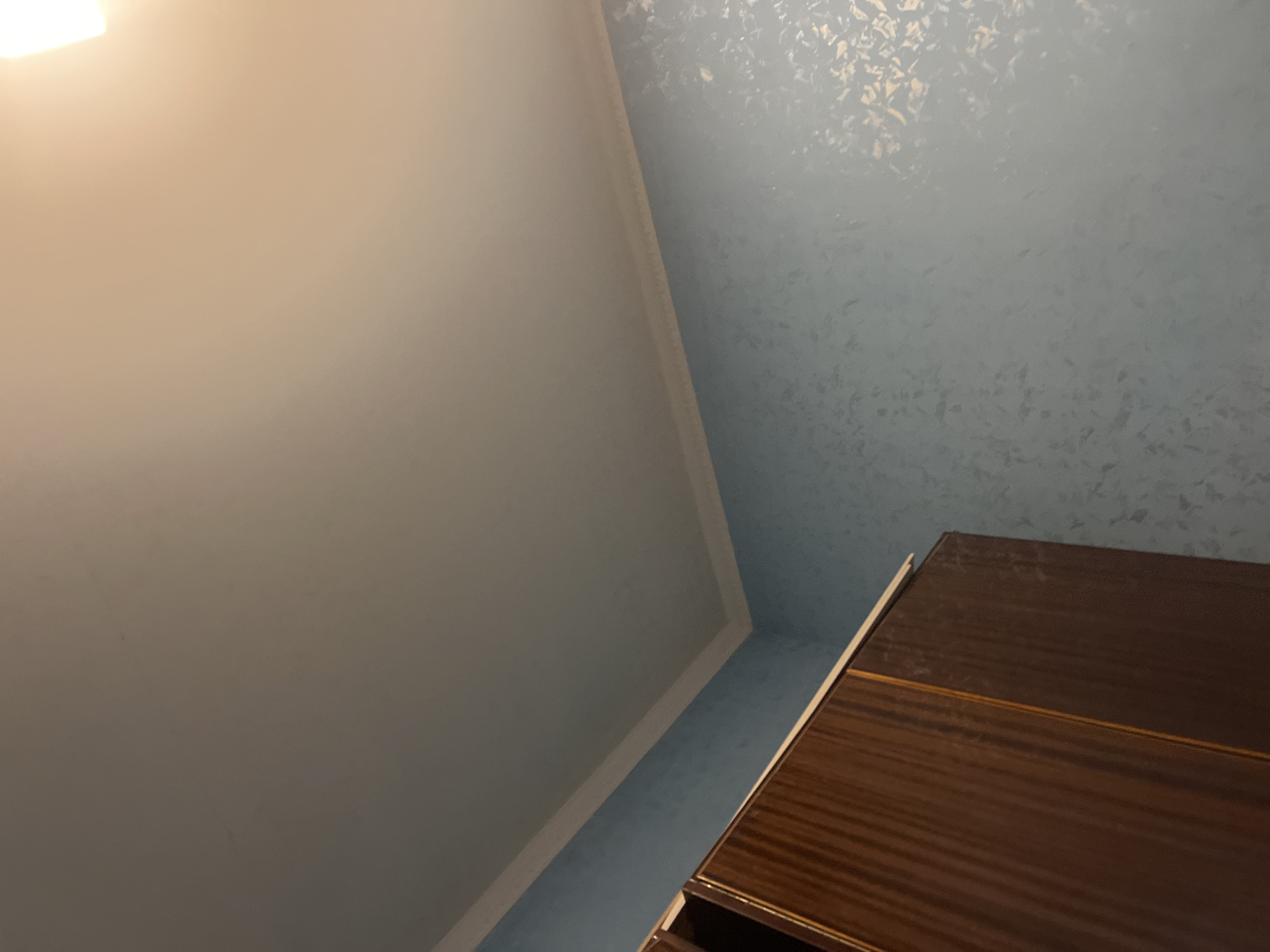 The image shows an interior view of a room, likely a bedroom, with a light blue textured wallpaper. A portion of a dark brown wooden wardrobe is visible on the left side of the frame, with a light-colored edge on top. The ceiling is off-white and there is white crown molding where the walls meet the ceiling. The lighting suggests it might be nighttime or that the room is dimly lit, with a bright light source, possibly a lamp, illuminating the upper right portion of the ceiling. There are no people or animals present in the image, and no specific actions are taking place. The scene depicts a static interior environment. The location in Jrarbi, Armenia is not visually discernible from the provided image.