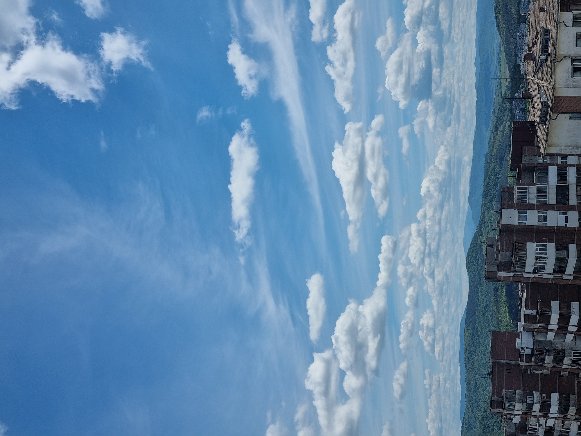 The image shows a view of Veliko Tarnovo, Bulgaria, with a clear blue sky filled with scattered white clouds. In the distance, a mountain range is visible, covered in green vegetation. Closer to the foreground, there are residential buildings with brick facades and windows, suggesting an urban area. The orientation of the image is vertical.