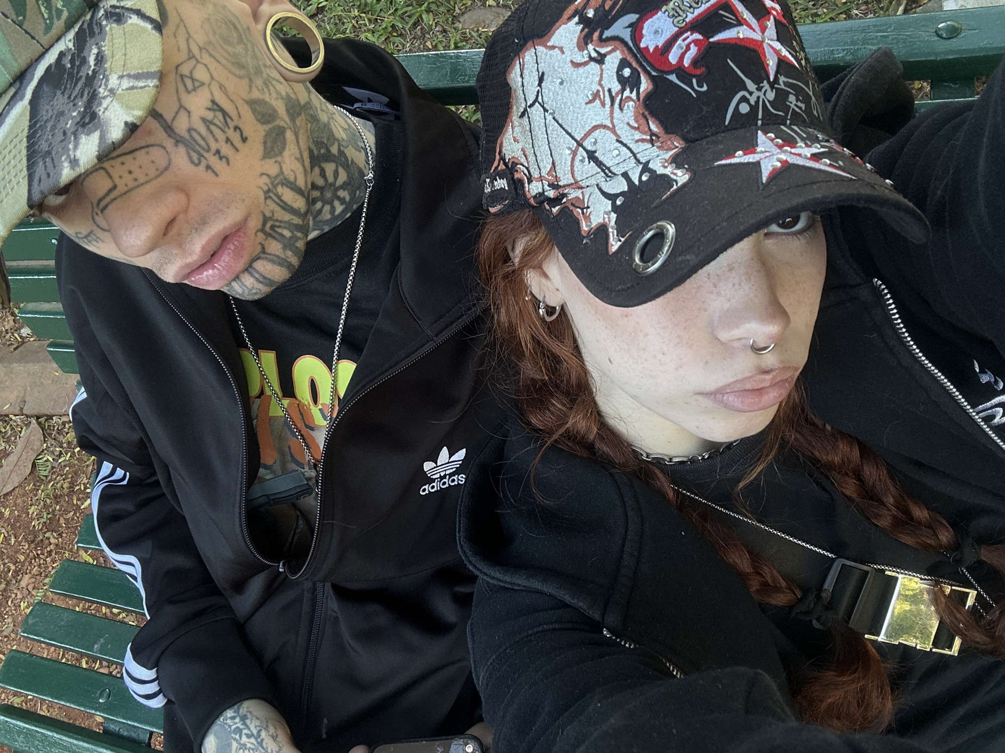 Two individuals are captured in a close-up, selfie-style photograph, seated side-by-side on a green slatted park bench, likely in Buenos Aires, Argentina. The setting appears to be outdoors during the daytime, with natural light and glimpses of foliage in the background.
The person on the left, with extensive facial and neck tattoos, wears a camouflage bucket hat, a large golden ear tunnel, and a black Adidas zip-up jacket with white stripes on the sleeves. Visible tattoos include a cartoon bandage on the nose and the numbers "1312" on the cheek. A silver chain necklace hangs around their neck.
The person on the right, with reddish-brown braided hair and noticeable freckles, wears a black cap adorned with red and white star designs and metal studs or grommets. They have a septum piercing and small hoop earrings. A silver chain necklace with a distinctive, rectangular pendant hangs over their black zip-up jacket. Their expression is neutral, with slightly pursed lips.
Both subjects are dressed in casual attire, suggesting a relaxed outing. A corner of a mobile phone is partially visible in the bottom right of the frame.