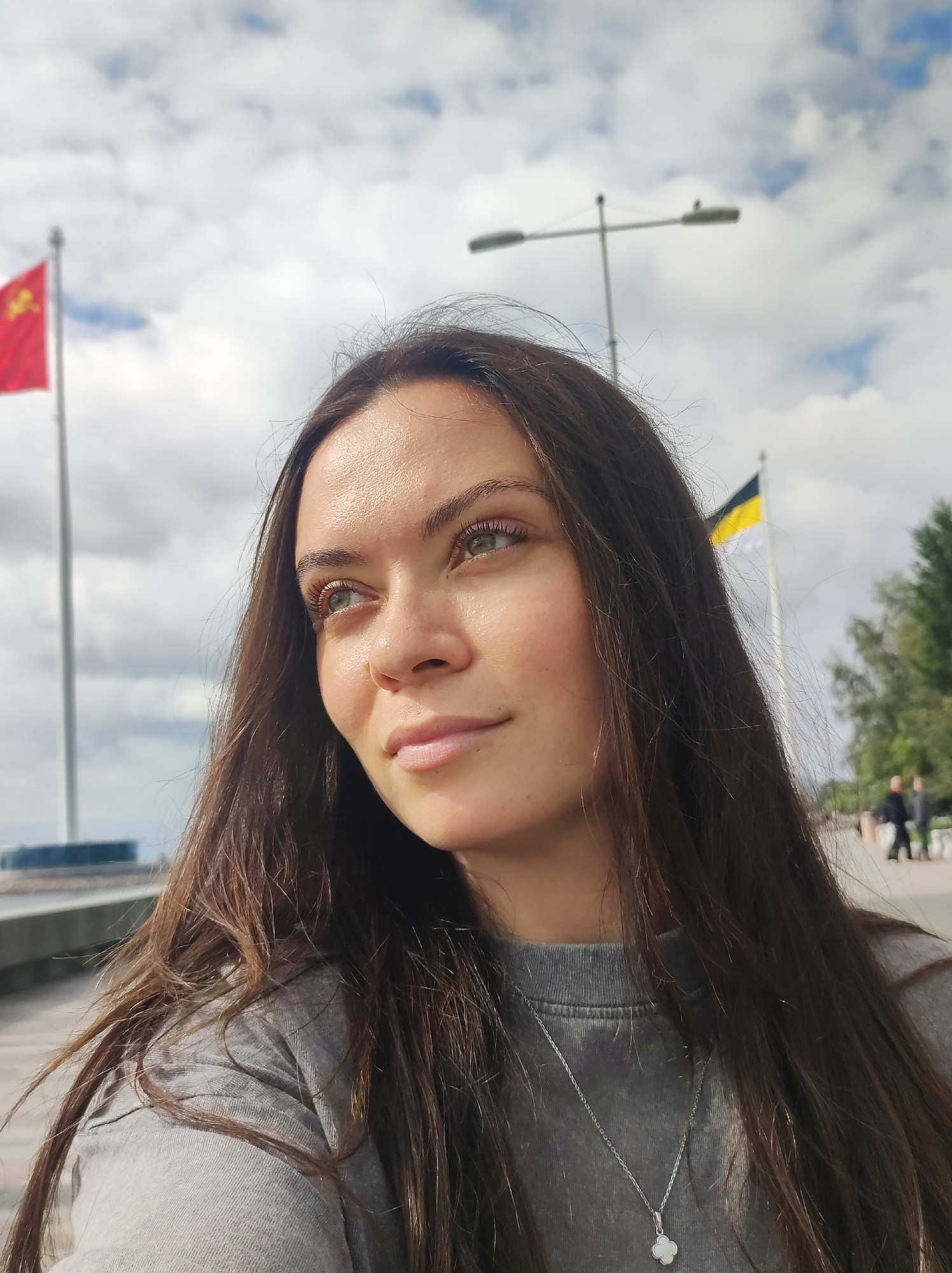 A woman with long, dark brown hair, wearing a grey sweatshirt and a silver pendant necklace, looks off to the left. She has light skin and green eyes. Behind her, a red flag with yellow symbols is visible on a flagpole, along with a street lamp. Further in the background, a black and yellow flag is also on a flagpole, with trees and people walking in the distance. The sky is cloudy. The scene appears to be outdoors, possibly along a waterfront or promenade, a common feature in Saint Petersburg, Russia.