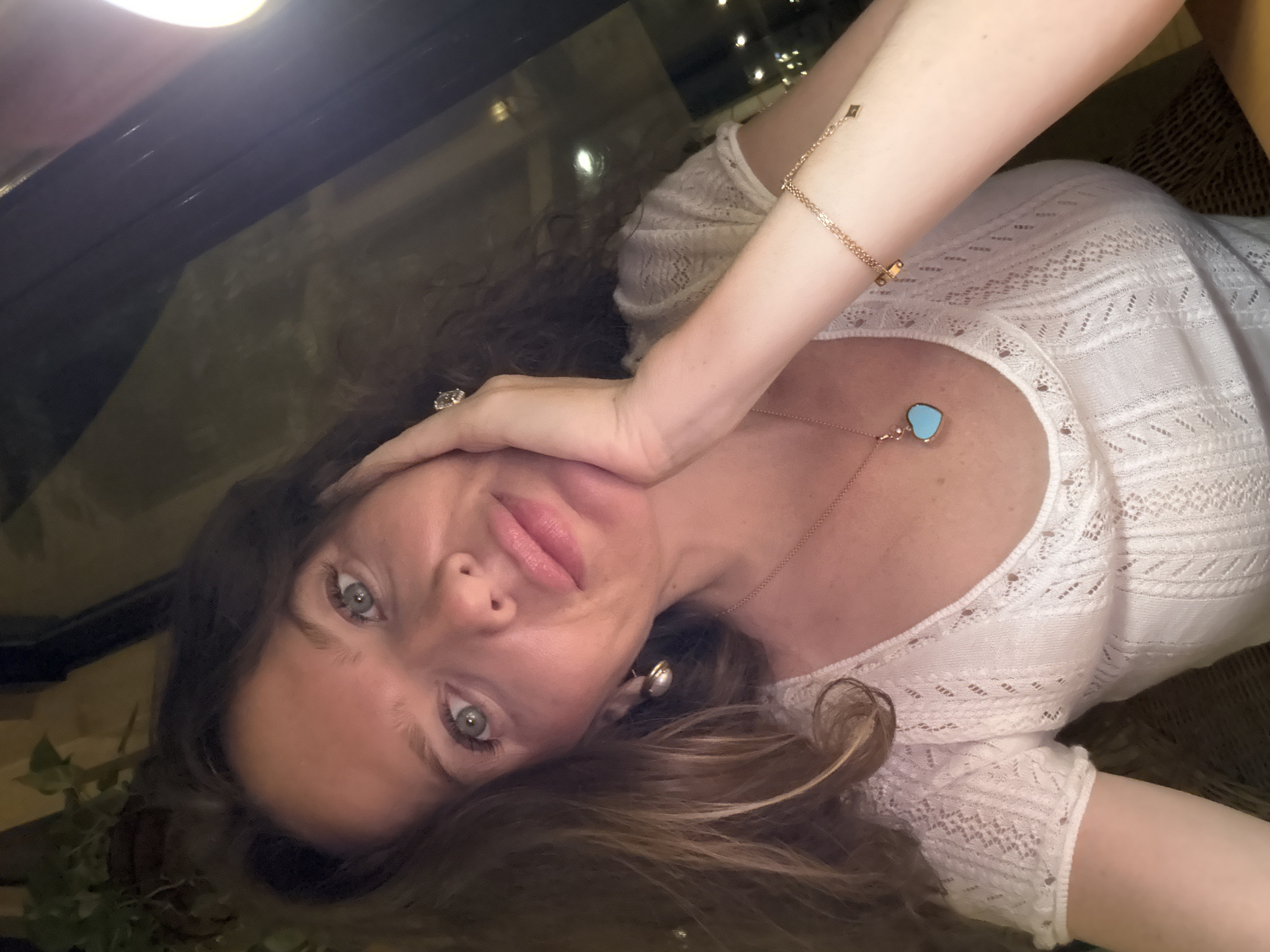 A selfie of a woman with long, dark, wavy hair and blue eyes is shown. She is wearing a white, knitted, short-sleeved top with a scoop neckline. She has on a gold necklace with a turquoise heart-shaped pendant and a gold bracelet with small square charms. Her left hand is resting on her cheek. The background is dark with some blurred lights, suggesting an indoor setting, possibly a restaurant or bar. No specific location in Dubai is identifiable.