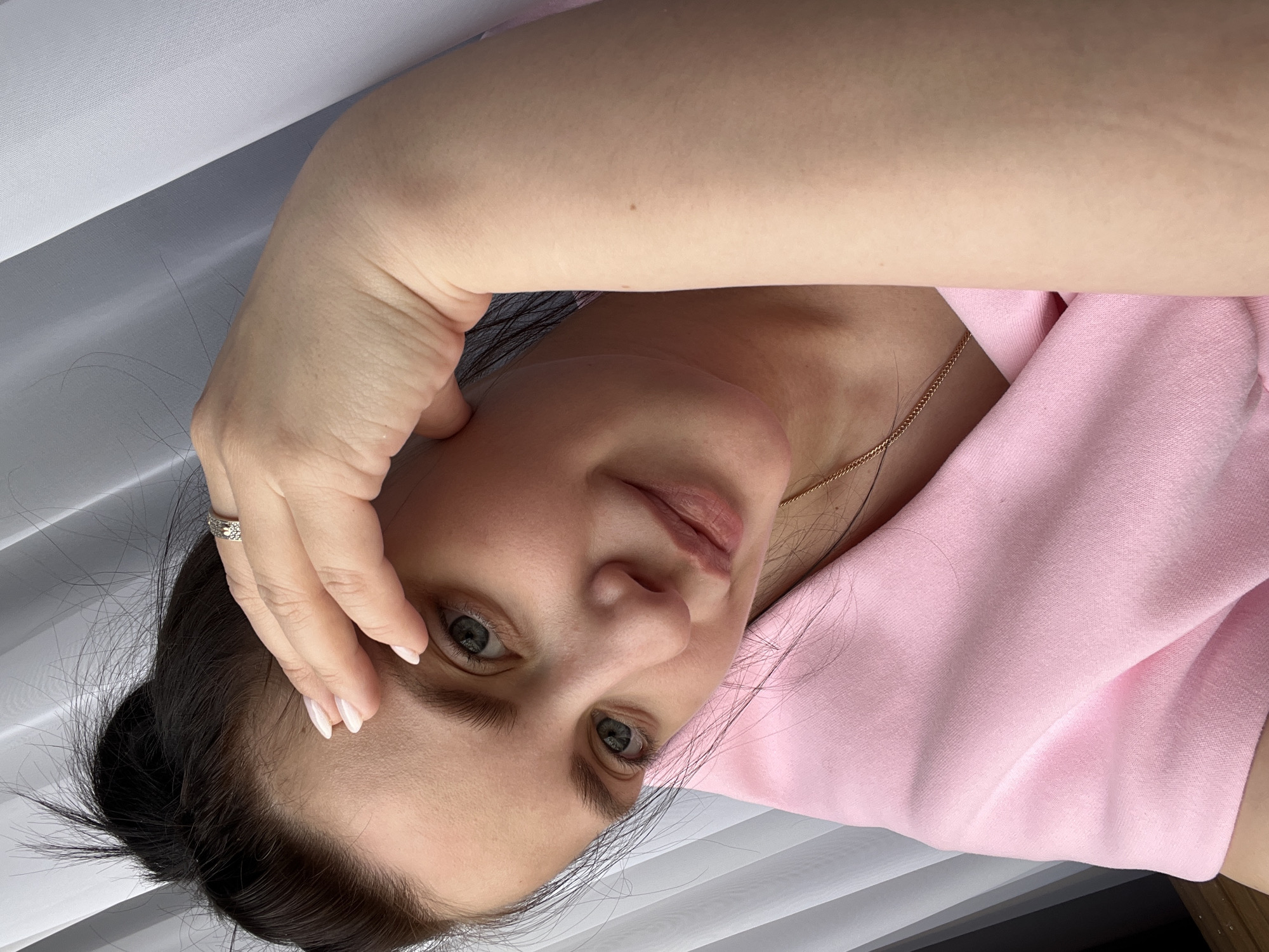A woman is posing for a selfie. She is wearing a pink top and a gold necklace. Her hair is dark and pulled back, and she is wearing a ring on her finger. She is holding her hand to her forehead and looking at the camera with a slight smile. The background is a blurred white surface. The location is Марковский, Russia.